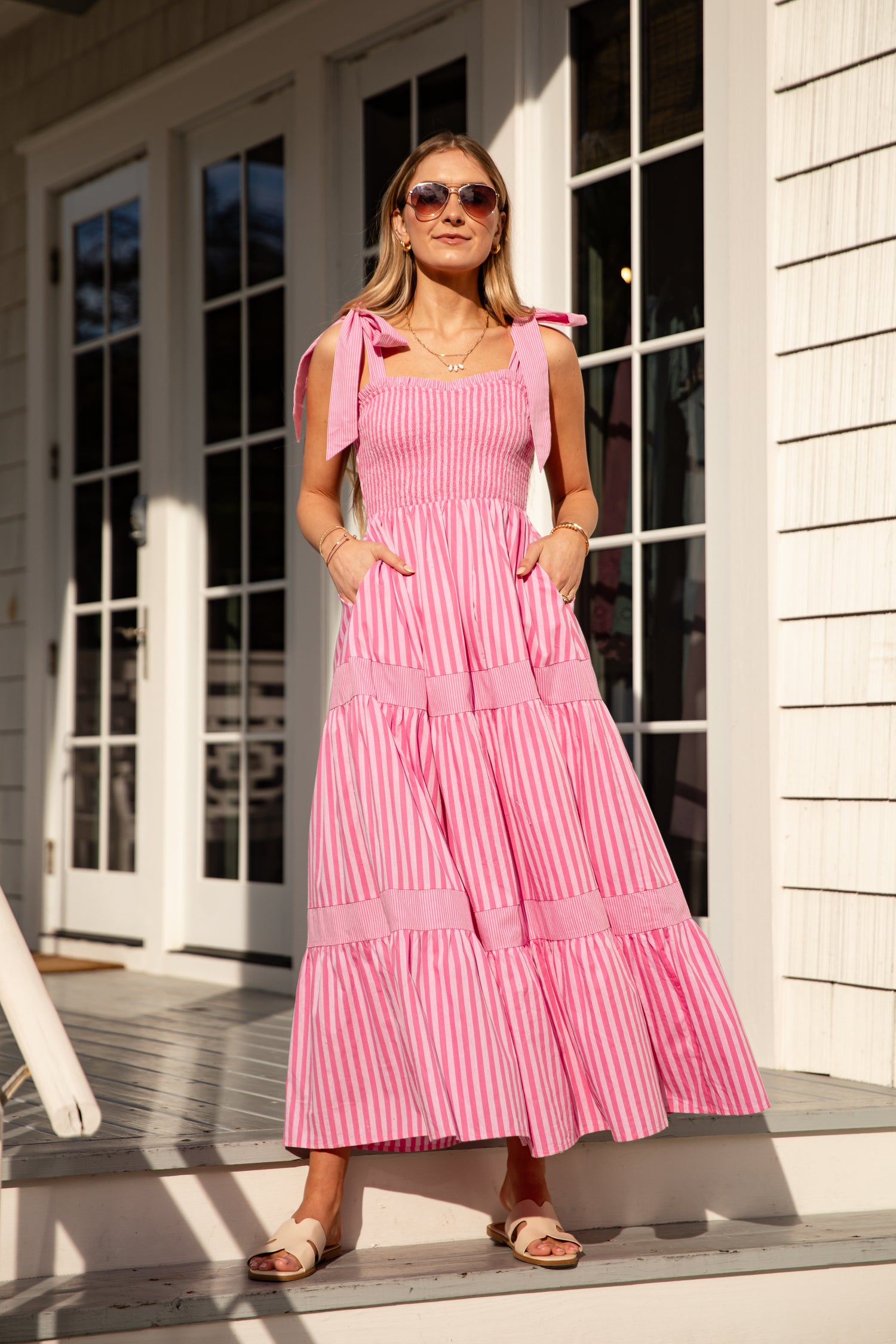 Woman wearing a pink striped dress with bow straps standing on a porch.