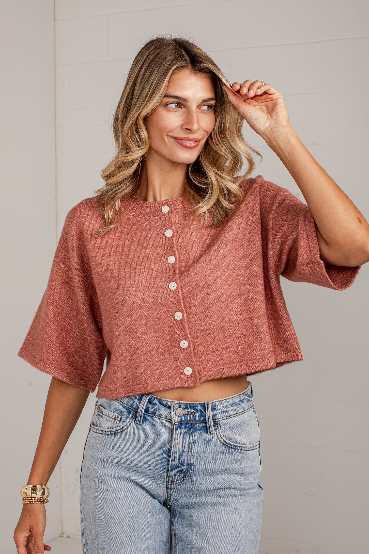A woman wearing a dusty rose short-sleeve cropped button-front top and light-wash blue jeans, standing against a light gray wall.
