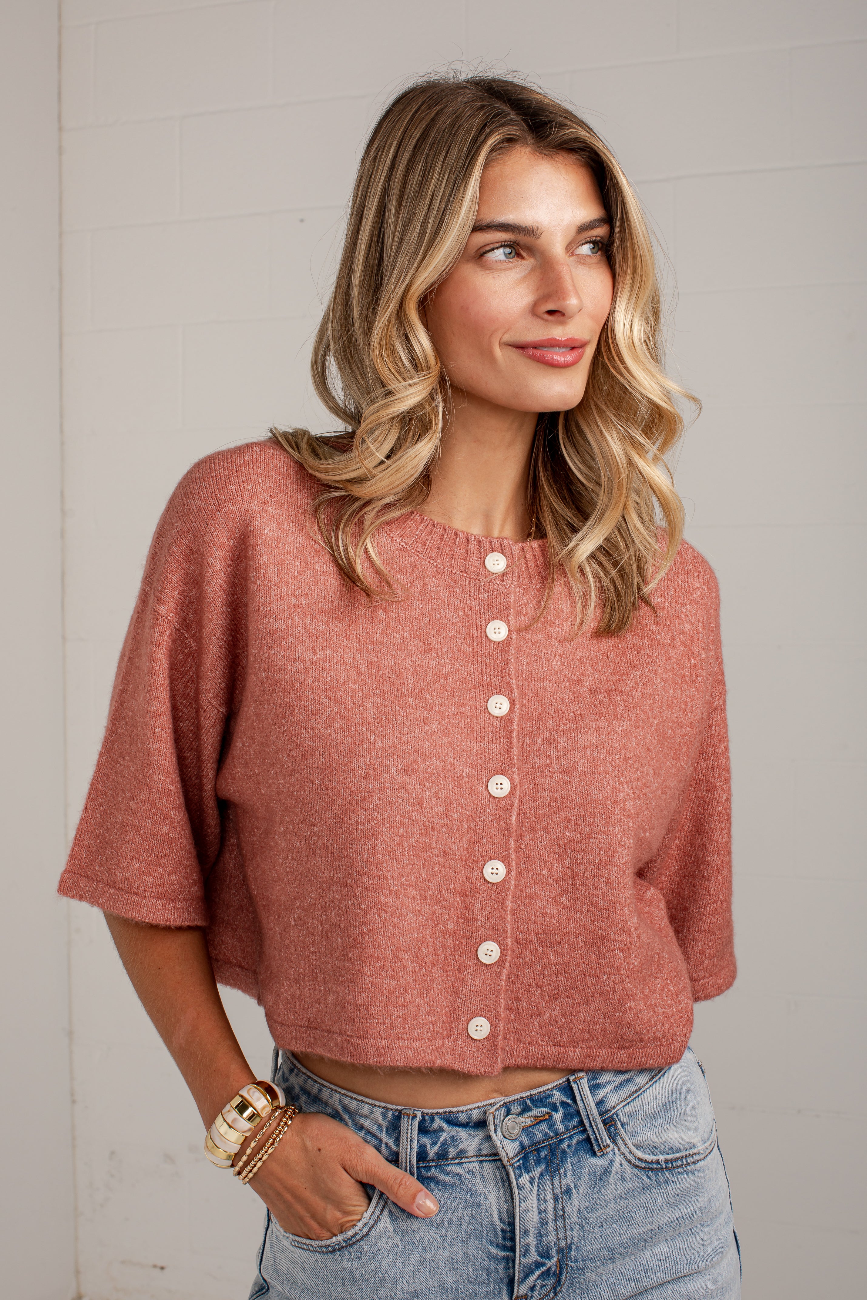 A woman wearing a dusty rose short-sleeve knit cardigan with white buttons, paired with light blue jeans, standing against a white brick wall.