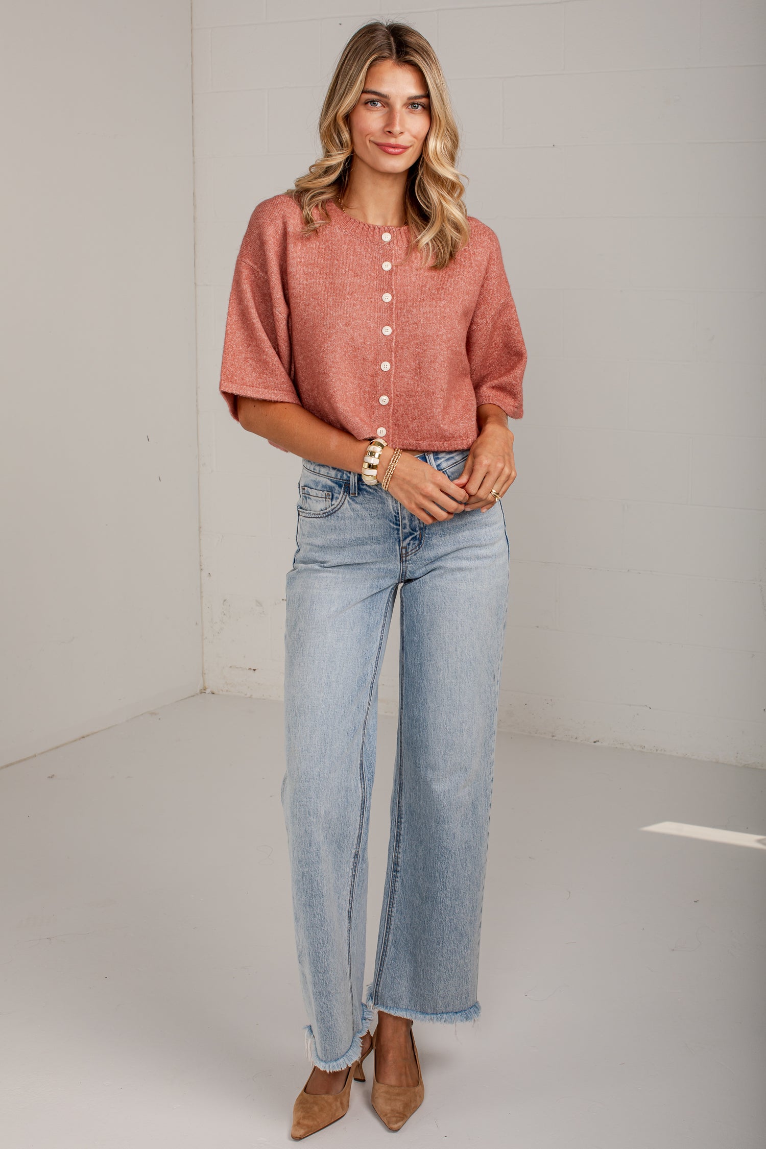 A woman modeling a short-sleeve, button-front top in a dusty rose color paired with light-wash wide-leg jeans and tan heels, standing against a plain white studio wall.