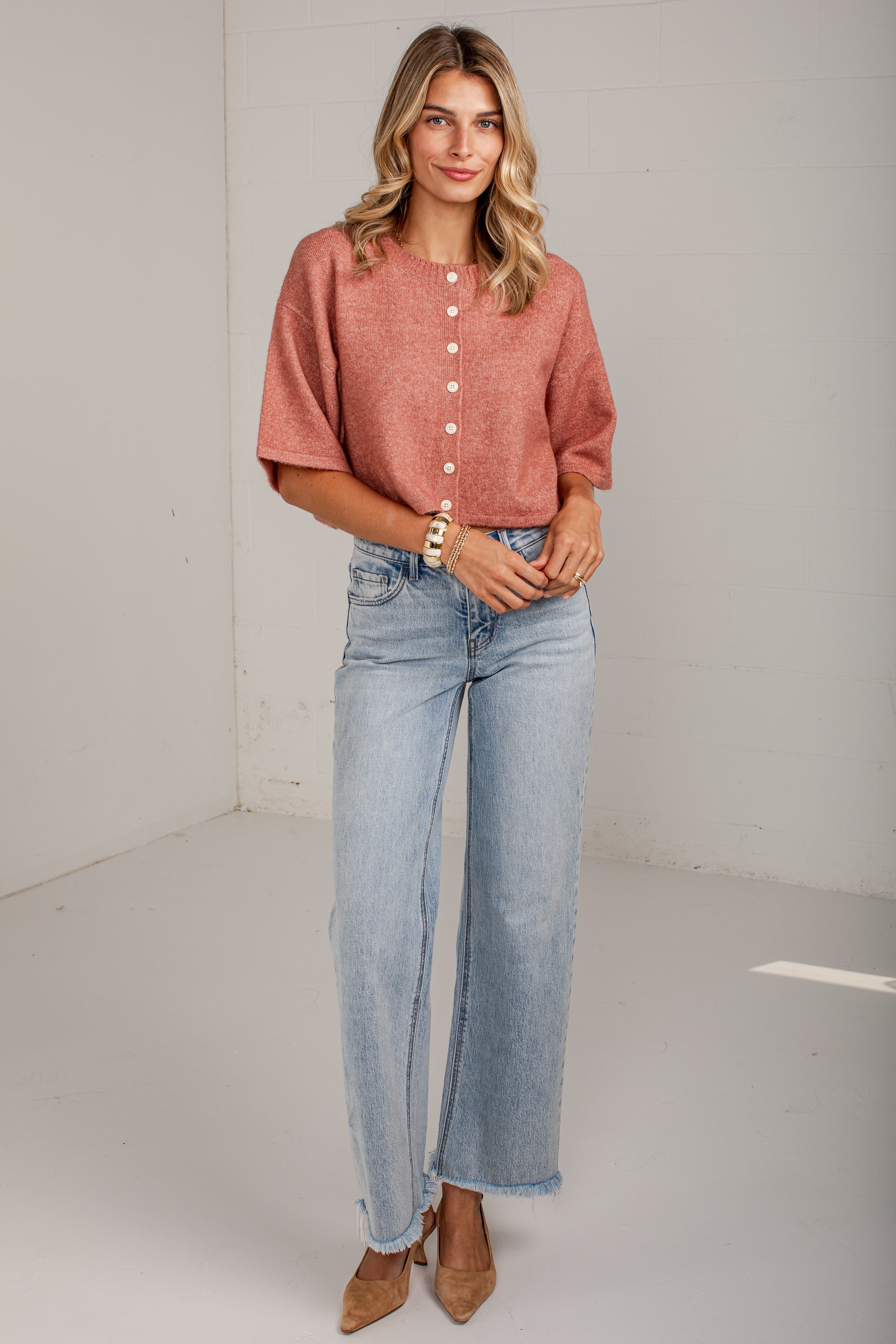 A woman modeling a short-sleeve, button-front top in a dusty rose color paired with light-wash wide-leg jeans and tan heels, standing against a plain white studio wall.