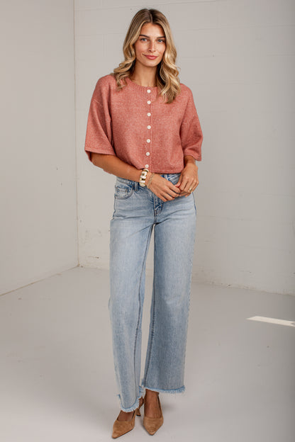 A woman modeling a short-sleeve, button-front top in a dusty rose color paired with light-wash wide-leg jeans and tan heels, standing against a plain white studio wall.