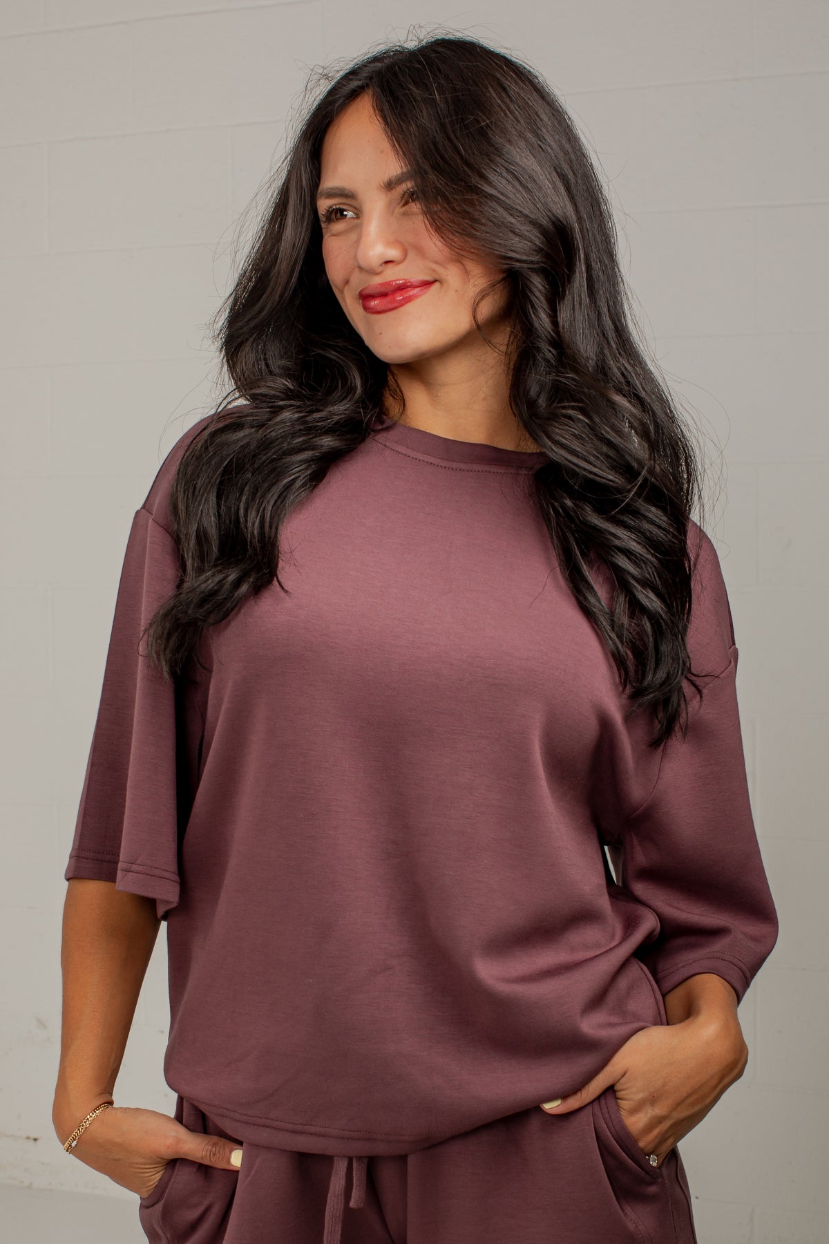 A woman with long, wavy dark hair wearing a matching dusty mauve lounge set, featuring a short-sleeve top and drawstring pants, posed against a light gray paneled wall.