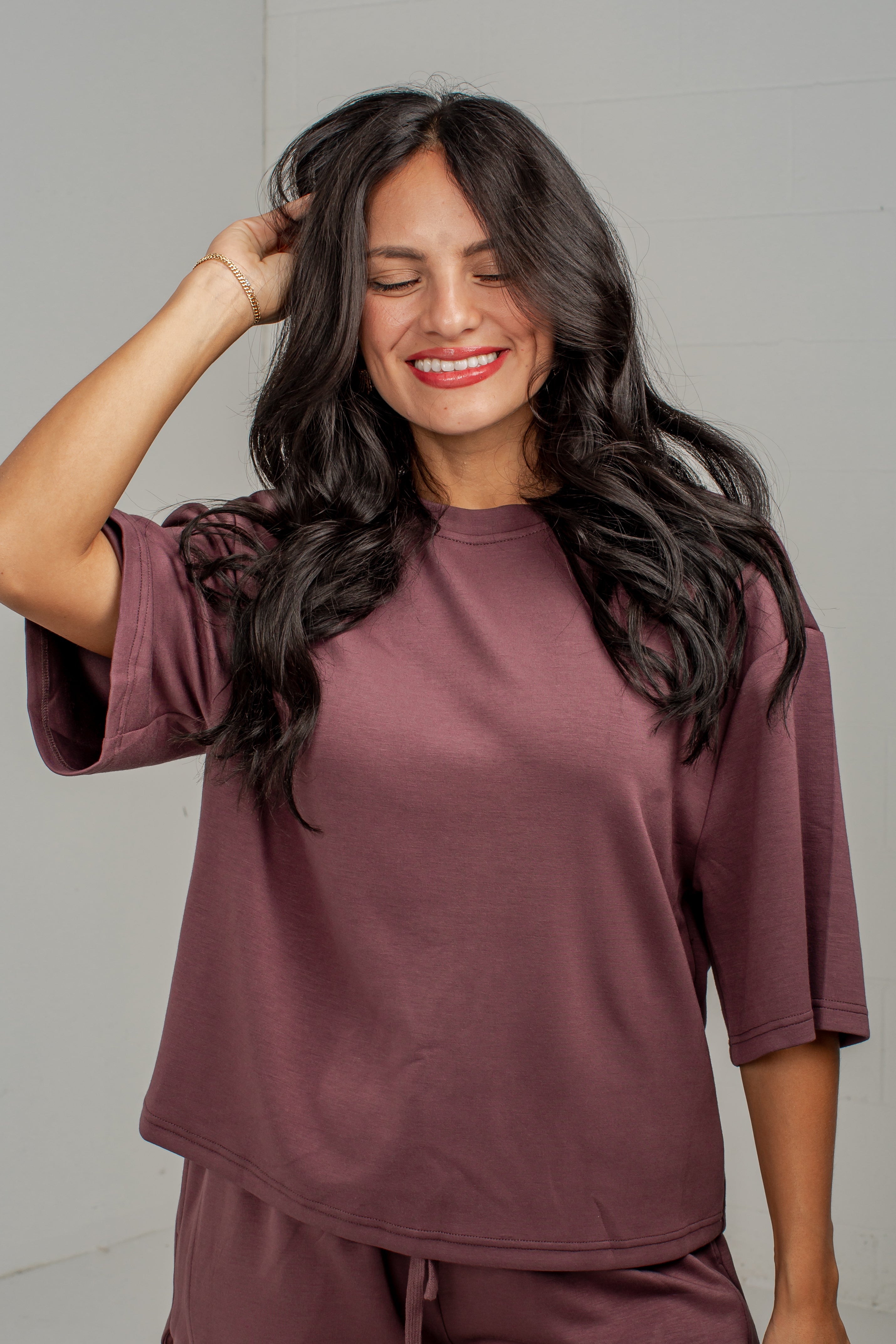 A woman with long, wavy dark hair is smiling and posing in a matching mauve-brown lounge set, featuring a short-sleeve top and pants, against a light gray wall.