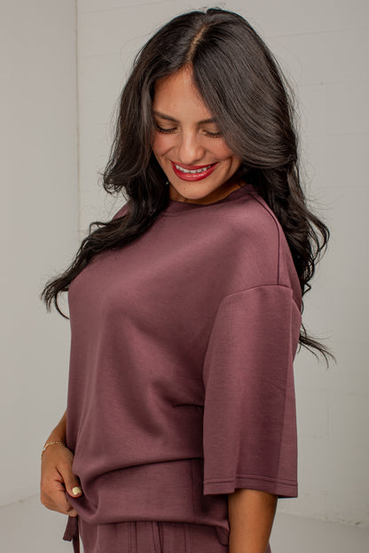 A woman wearing a dusty rose mauve short-sleeve top and matching bottoms, smiling and looking down against a plain light gray background.