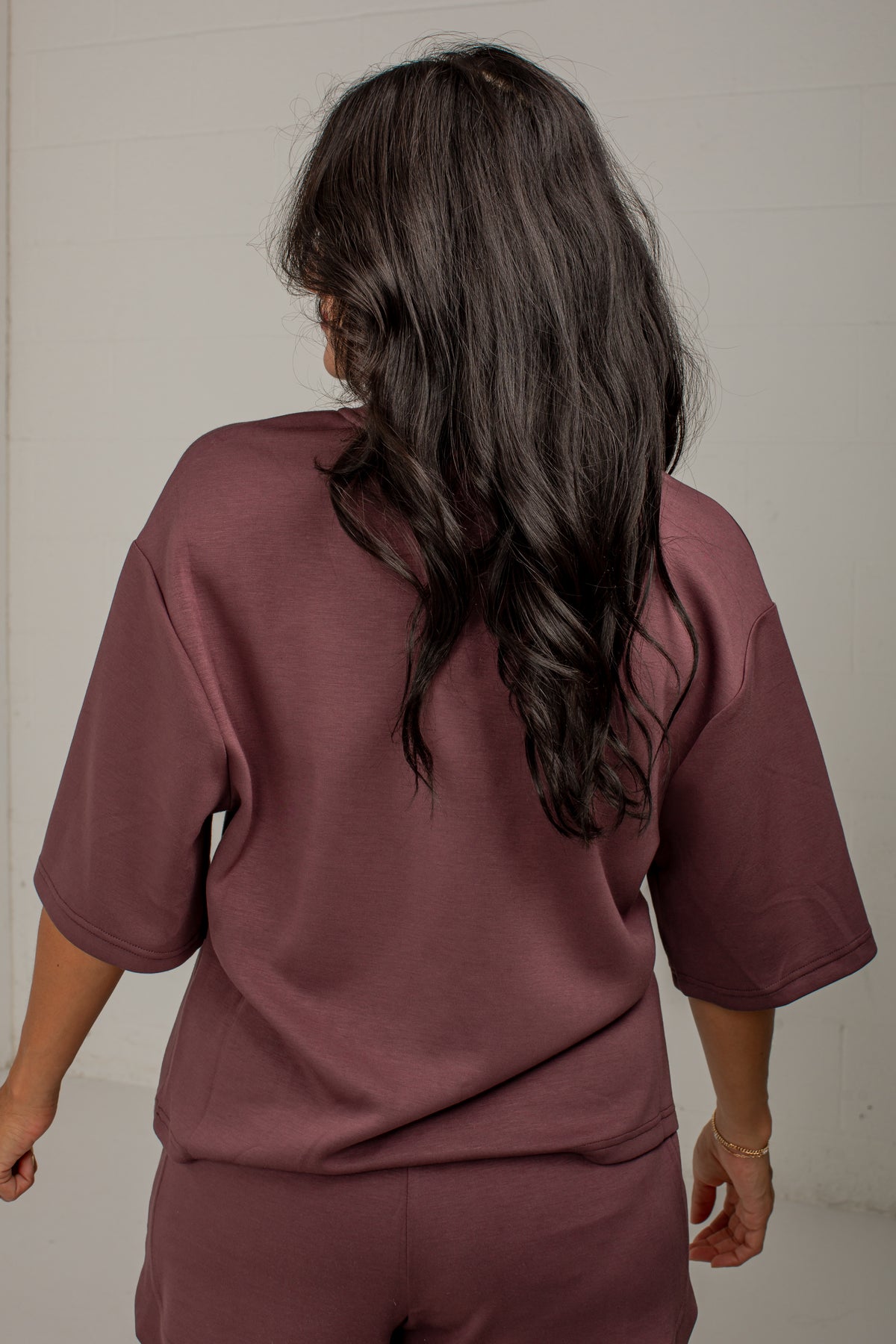 A woman viewed from behind wearing a matching mauve-brown short-sleeve top and shorts set against a plain off-white wall.