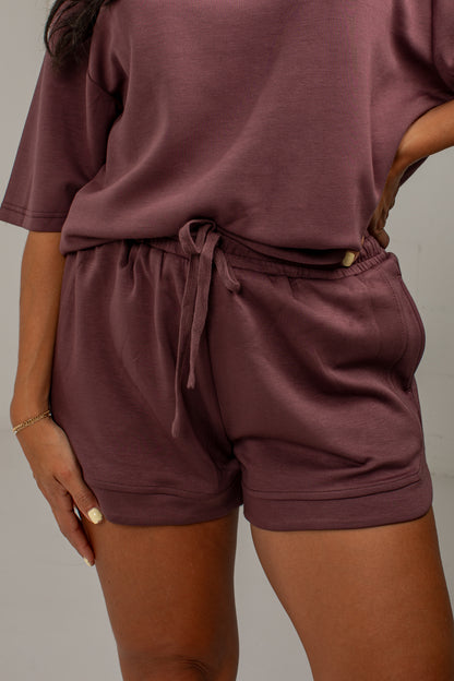 A close-up of a person wearing a matching mauve lounge set, featuring a short-sleeve top and drawstring shorts, against a plain light gray background.