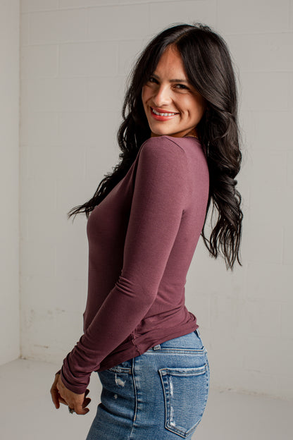 A woman with long dark hair is shown from the back and side, wearing a fitted long-sleeve top in a muted plum-brown color and blue jeans, standing against a plain light wall.