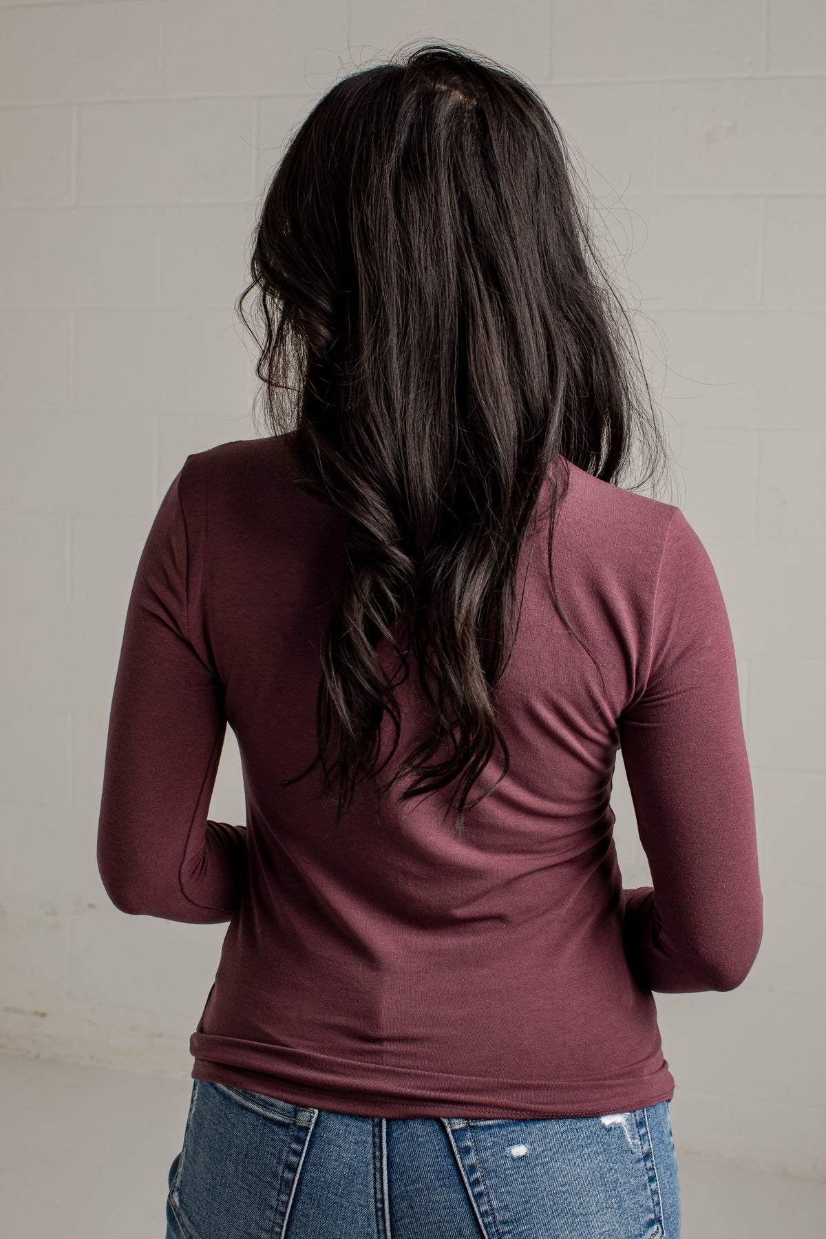 A woman viewed from behind wearing a fitted long-sleeve burgundy top and blue jeans against a light gray brick wall.