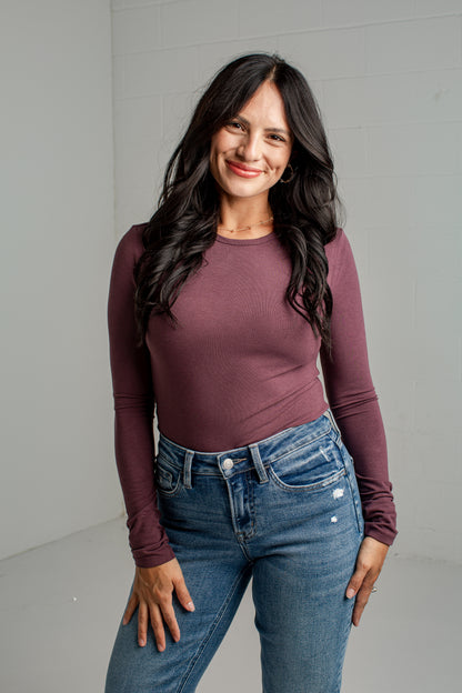 A woman with long dark hair wearing a fitted long-sleeve crewneck top in a dusty plum purple and blue jeans, standing against a light gray wall.