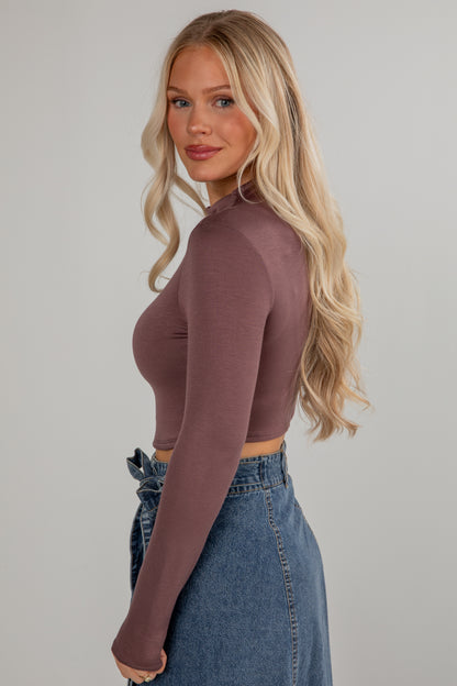Woman wearing a brown long-sleeve top and blue jeans against a plain background