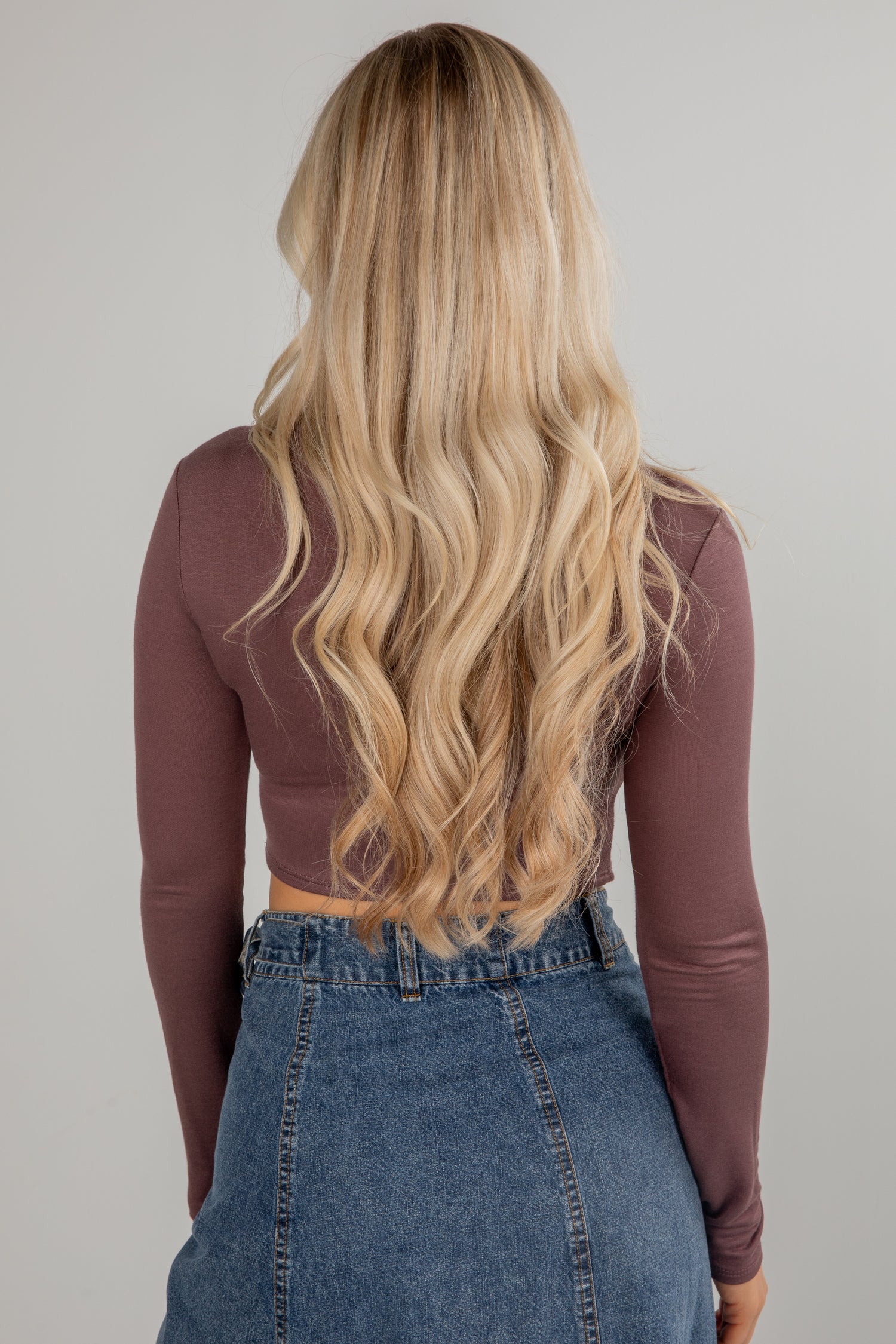 Person with long blonde hair wearing a brown long-sleeve top and blue jeans on a plain background