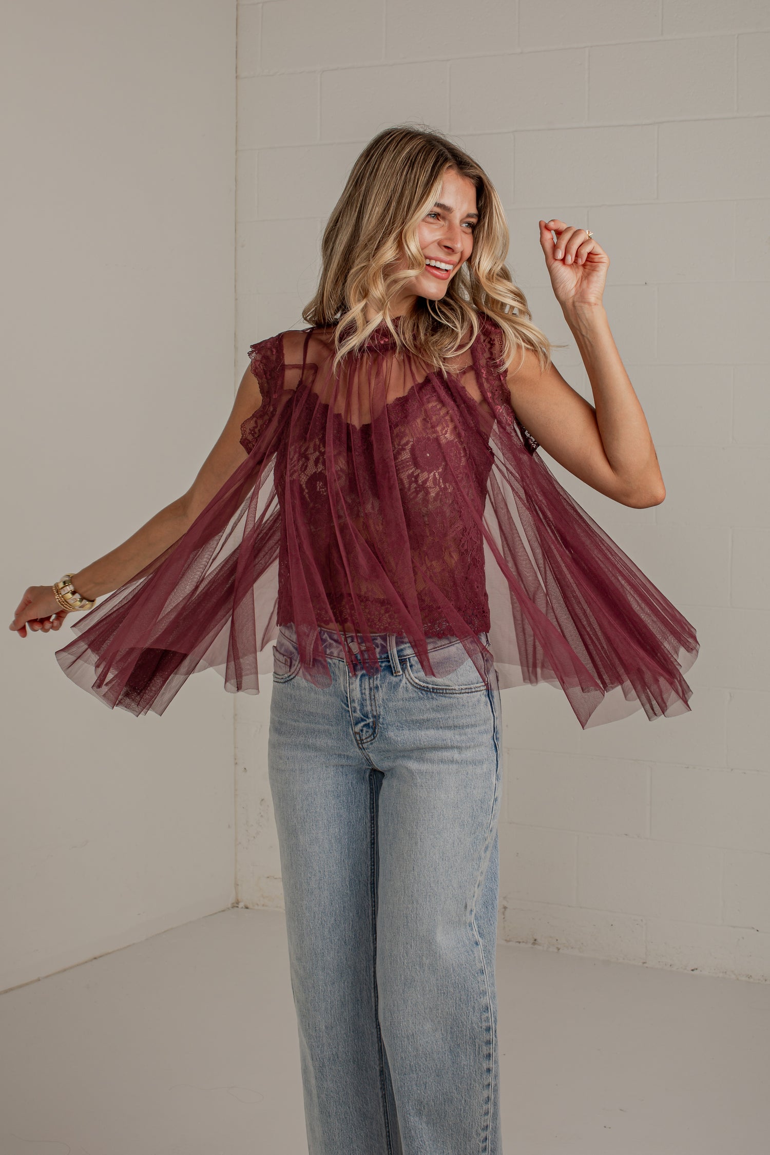 A woman modeling a sleeveless burgundy tulle top with sheer mesh panels and a floral lace overlay, paired with light-wash blue jeans in a bright indoor setting.