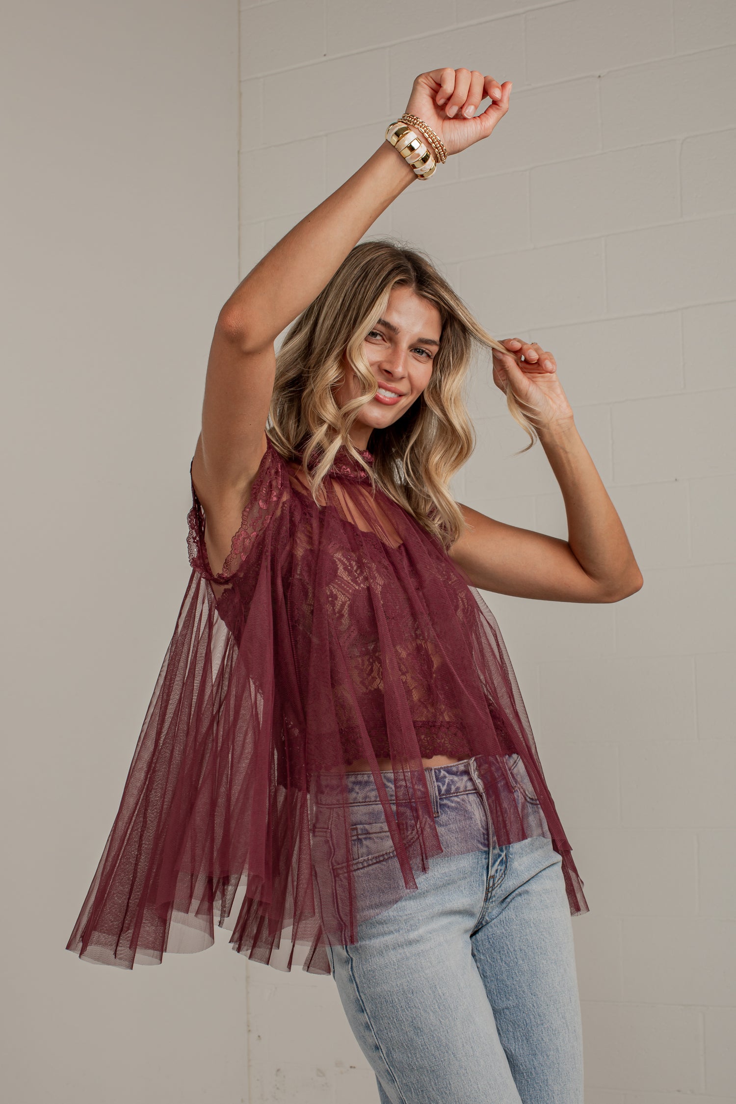 A woman modeling a sleeveless burgundy tulle top with a sheer lace overlay and flared tulle skirt, paired with light blue jeans, posing against a light gray brick wall.