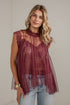 A woman wearing a sheer burgundy lace and tulle sleeveless top with a high mock neck, paired with light blue jeans, standing against a white brick wall.