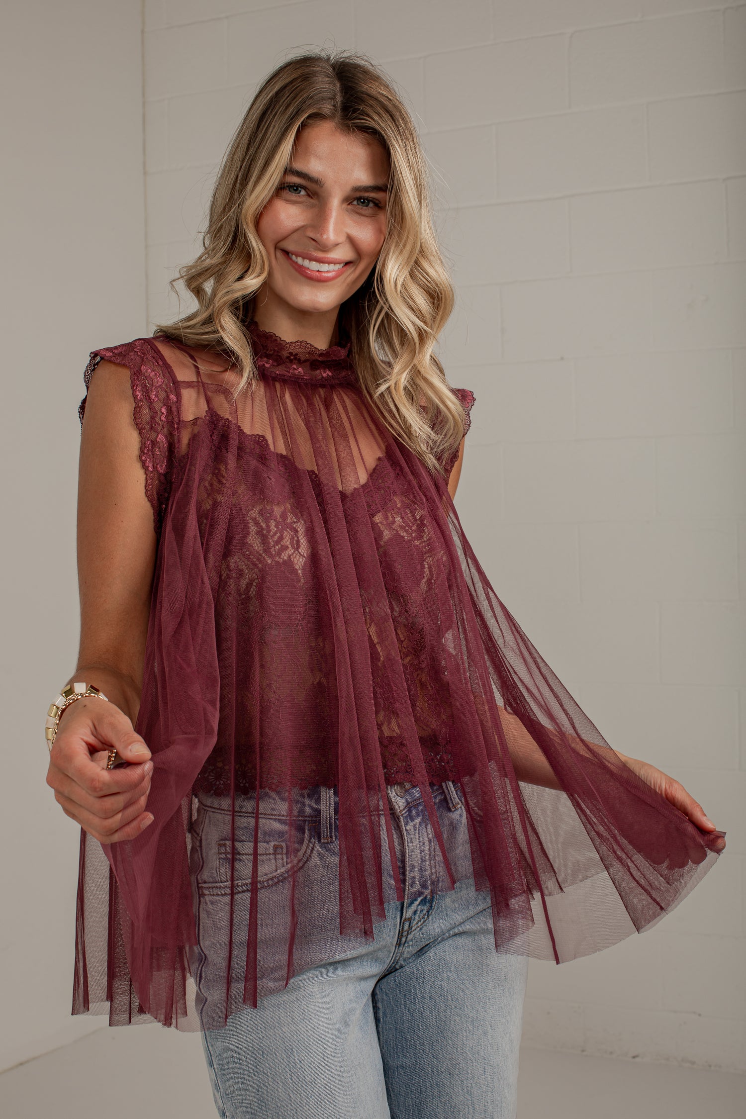 A woman wearing a sheer burgundy lace and tulle sleeveless top with light blue jeans, standing against a white brick wall.