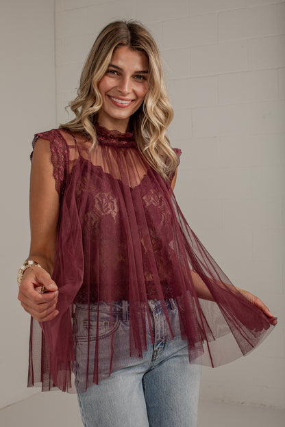 A woman wearing a sheer burgundy lace and tulle sleeveless top with light blue jeans, standing against a white brick wall.