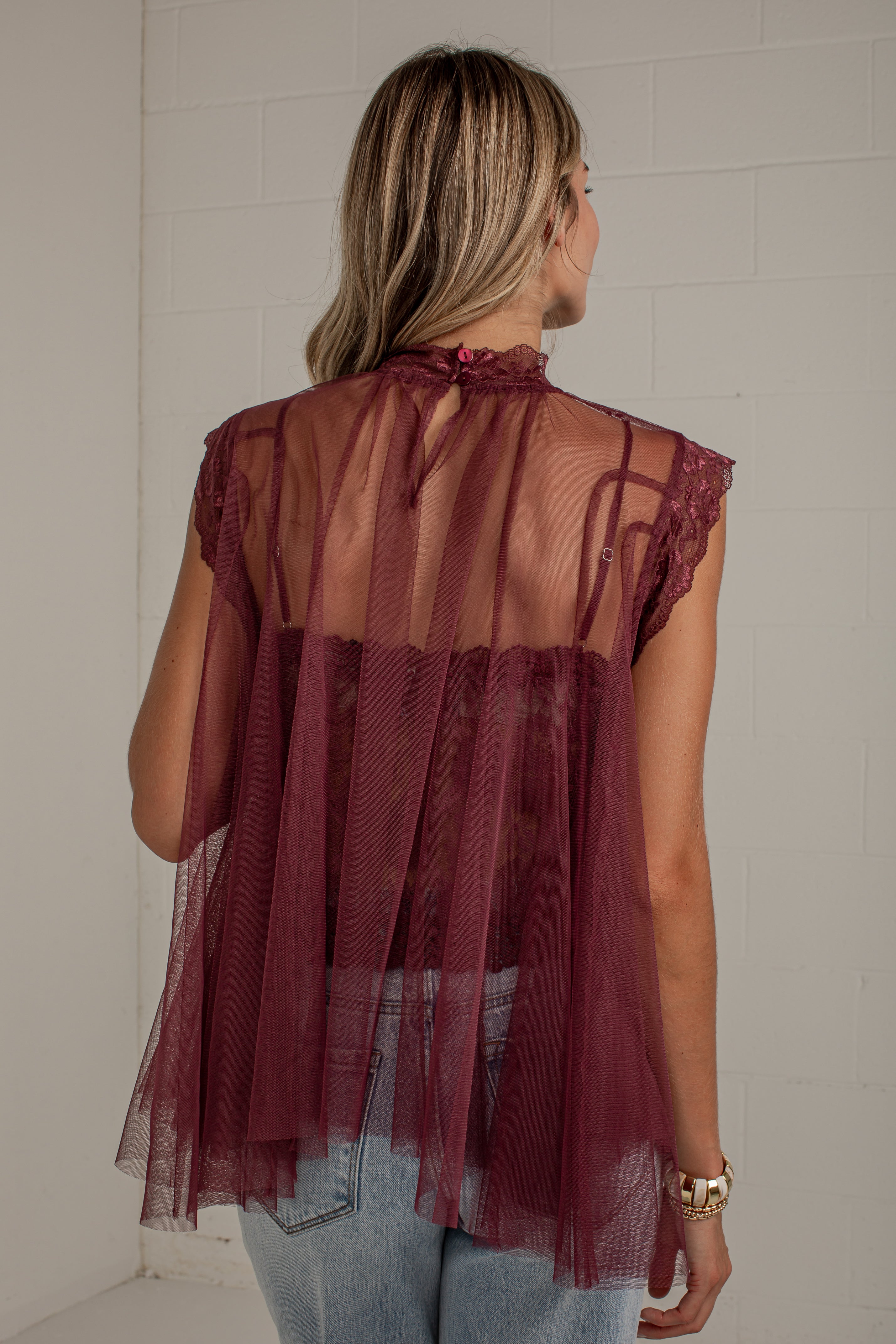 A woman viewed from behind wearing a sheer burgundy tulle top with lace trim over a matching camisole and light blue jeans, standing against a white wall.
