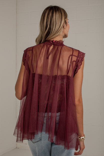 A woman viewed from behind wearing a sheer burgundy tulle top with lace trim over a matching camisole and light blue jeans, standing against a white wall.