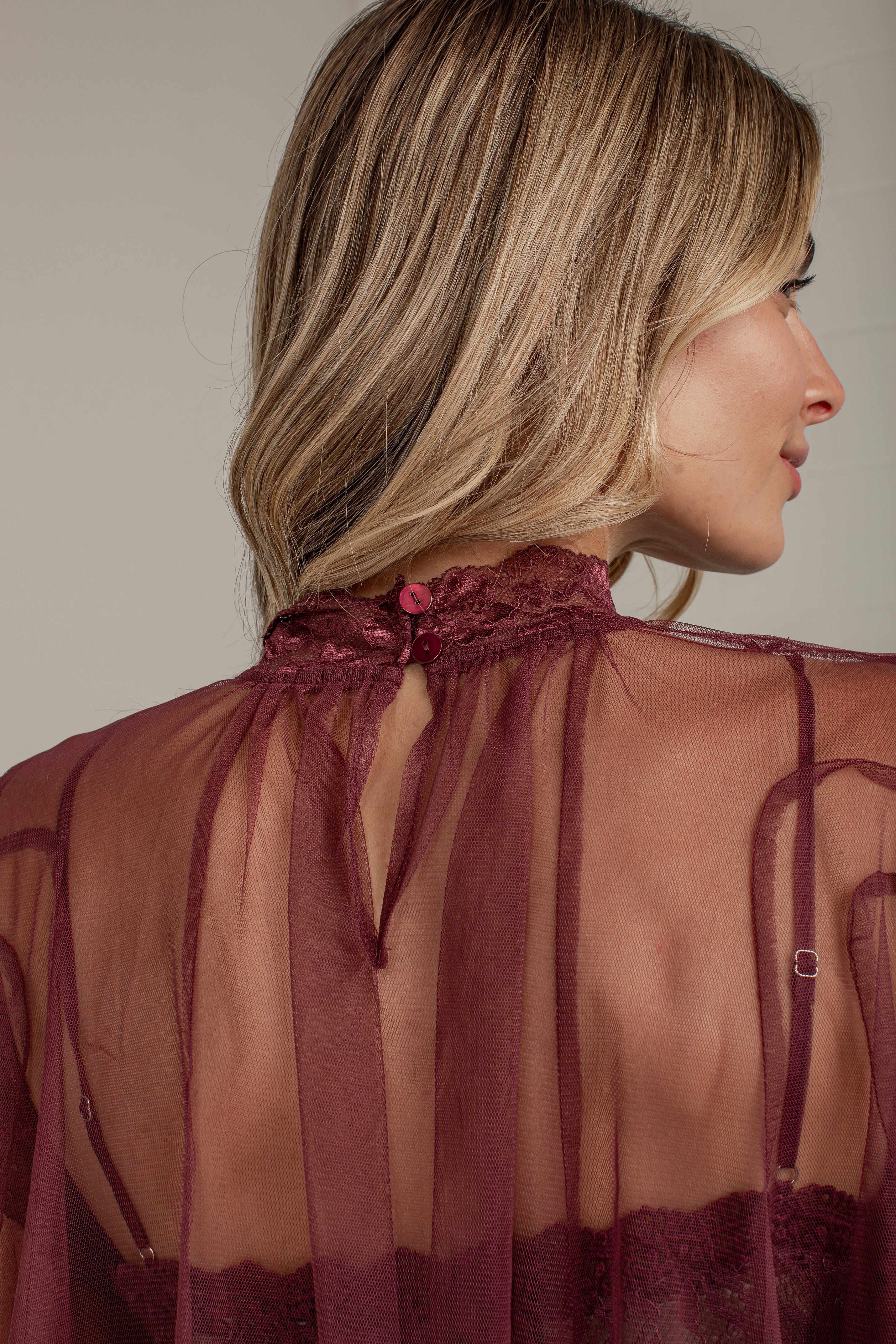 A woman shown from the back wearing a sheer burgundy mesh top with lace trim and tie details at the high neckline, photographed against a plain light gray studio background.
