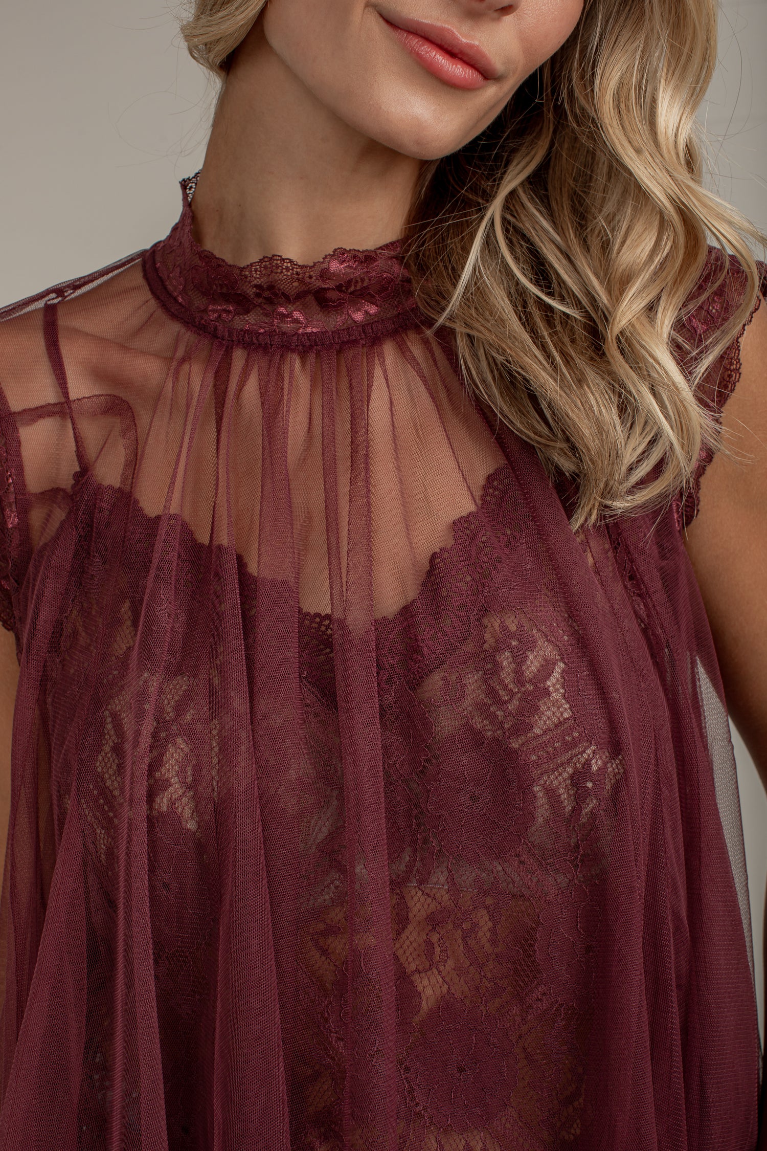 A close-up of a woman wearing a sheer burgundy lace and mesh top with a high ruffled neckline and floral lace pattern.