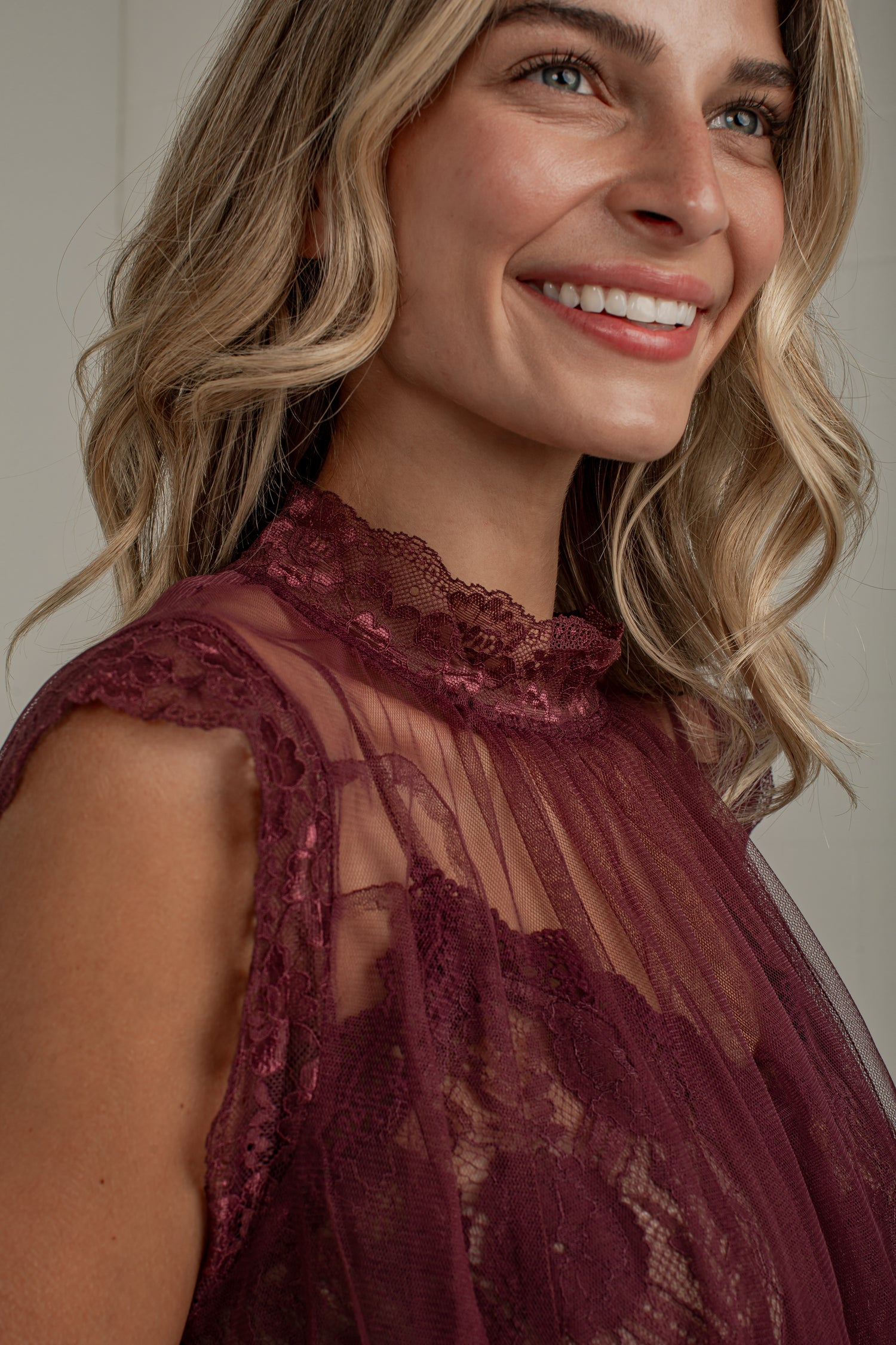 A close-up of a smiling woman with blonde hair wearing a sheer burgundy lace top with a high ruffled neckline.