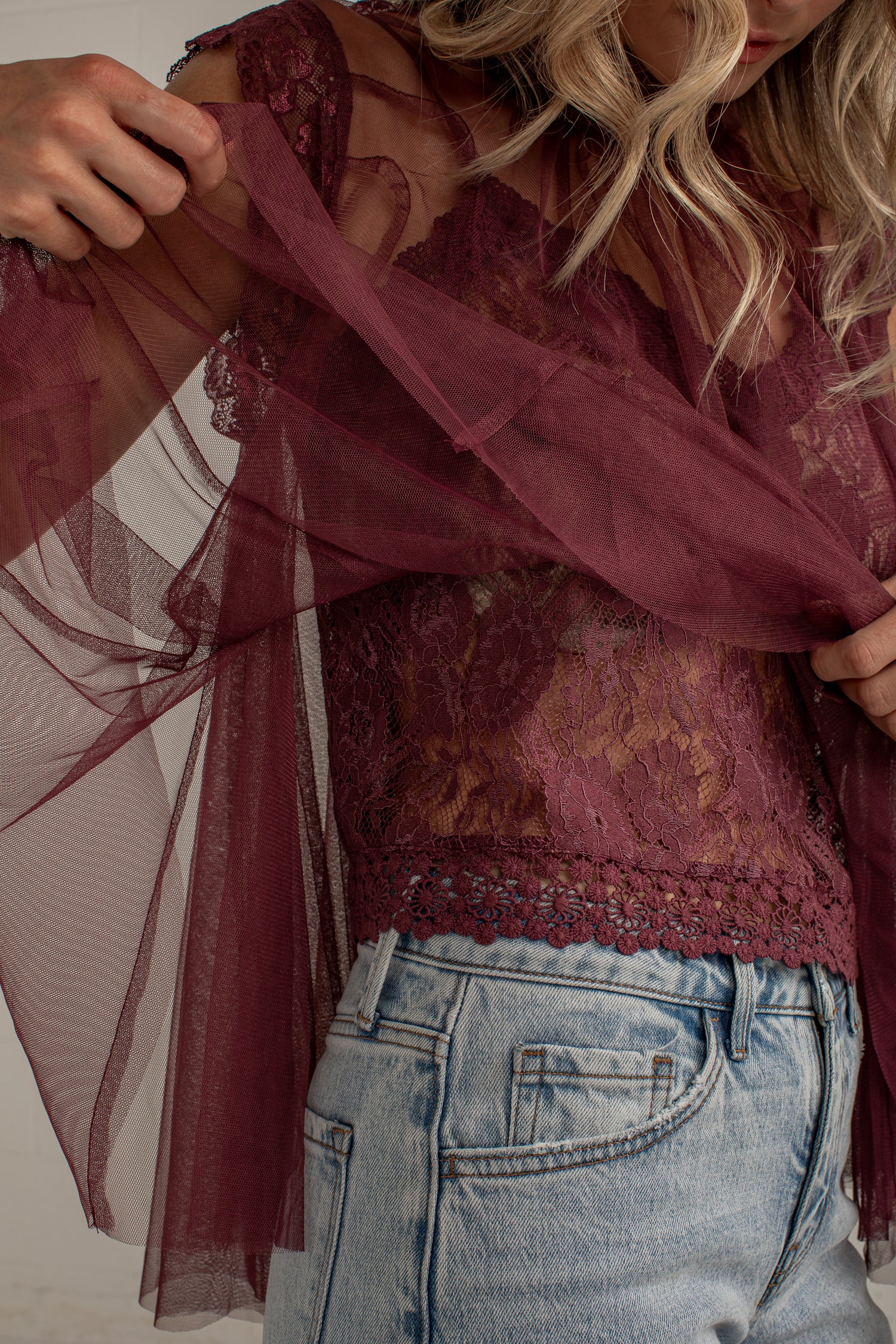 A close-up of a woman wearing a sheer burgundy tulle overlay and matching lace camisole top, paired with light blue jeans.