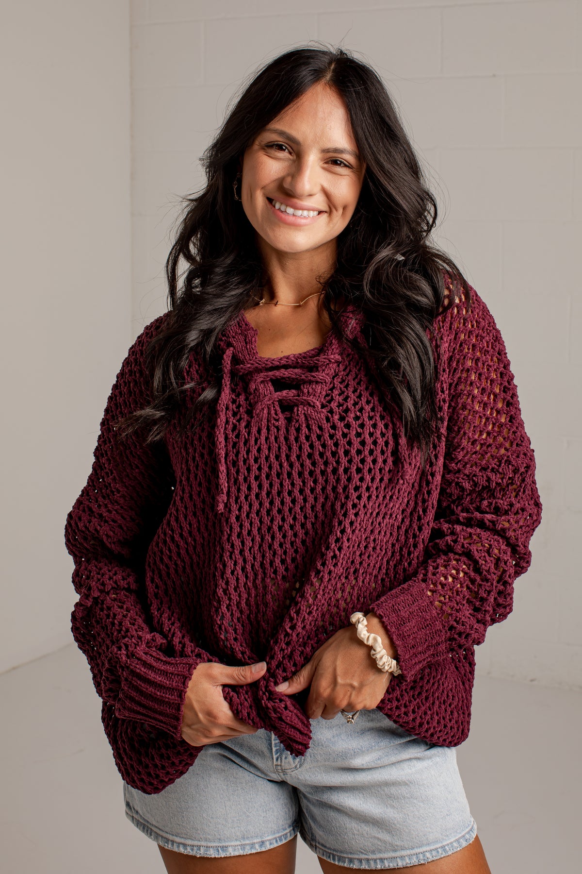 A smiling woman with long dark hair wears an oversized chunky-knit maroon sweater with tie details at the neckline, paired with light-wash denim shorts, standing against a light gray wall.
