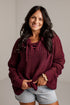 A smiling woman with long dark hair wears an oversized chunky-knit maroon sweater with tie details at the neckline, paired with light-wash denim shorts, standing against a light gray wall.