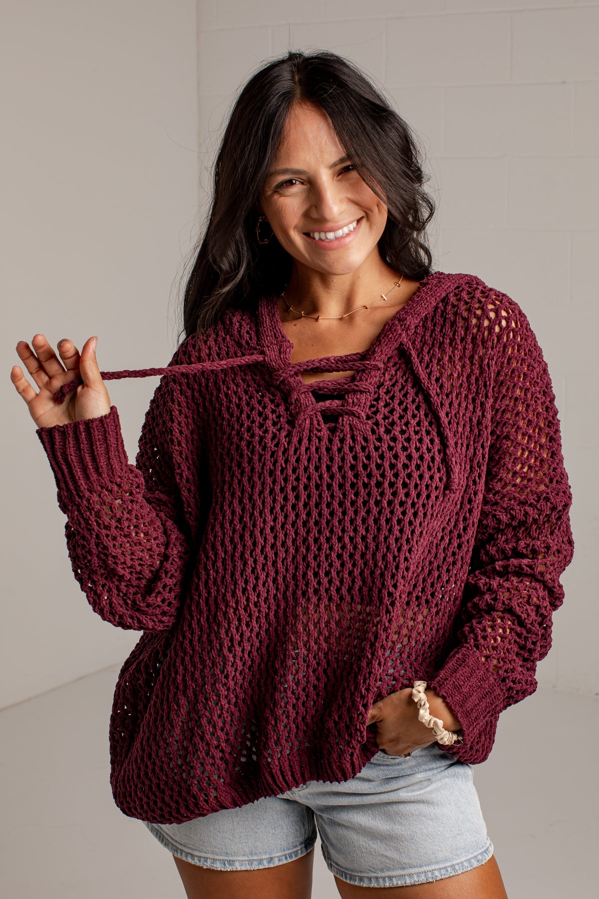 A woman wearing a chunky knit burgundy sweater with a lace-up neckline and light denim shorts, standing against a light gray wall.