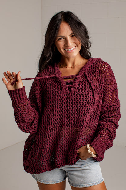 A woman wearing a chunky knit burgundy sweater with a lace-up neckline and light denim shorts, standing against a light gray wall.
