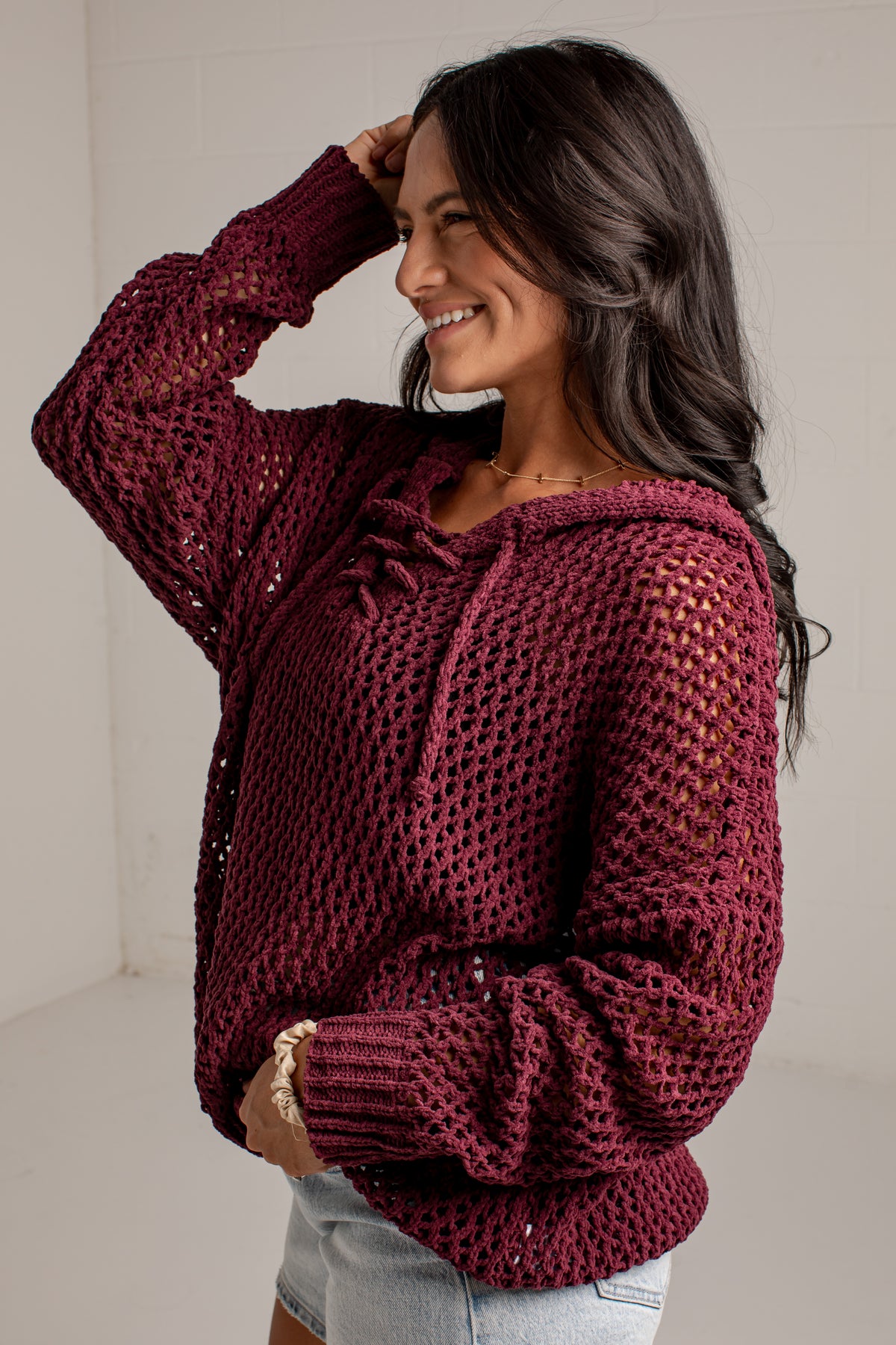 A woman wearing a chunky knit maroon open-knit hoodie sweater with drawstrings, paired with light-wash denim shorts, in a bright indoor setting.