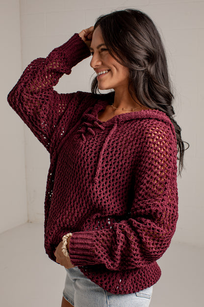 A woman wearing a chunky knit maroon open-knit hoodie sweater with drawstrings, paired with light-wash denim shorts, in a bright indoor setting.