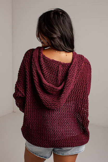 A woman viewed from behind wearing a loose, open-knit burgundy hooded sweater and denim shorts against a plain light gray wall.
