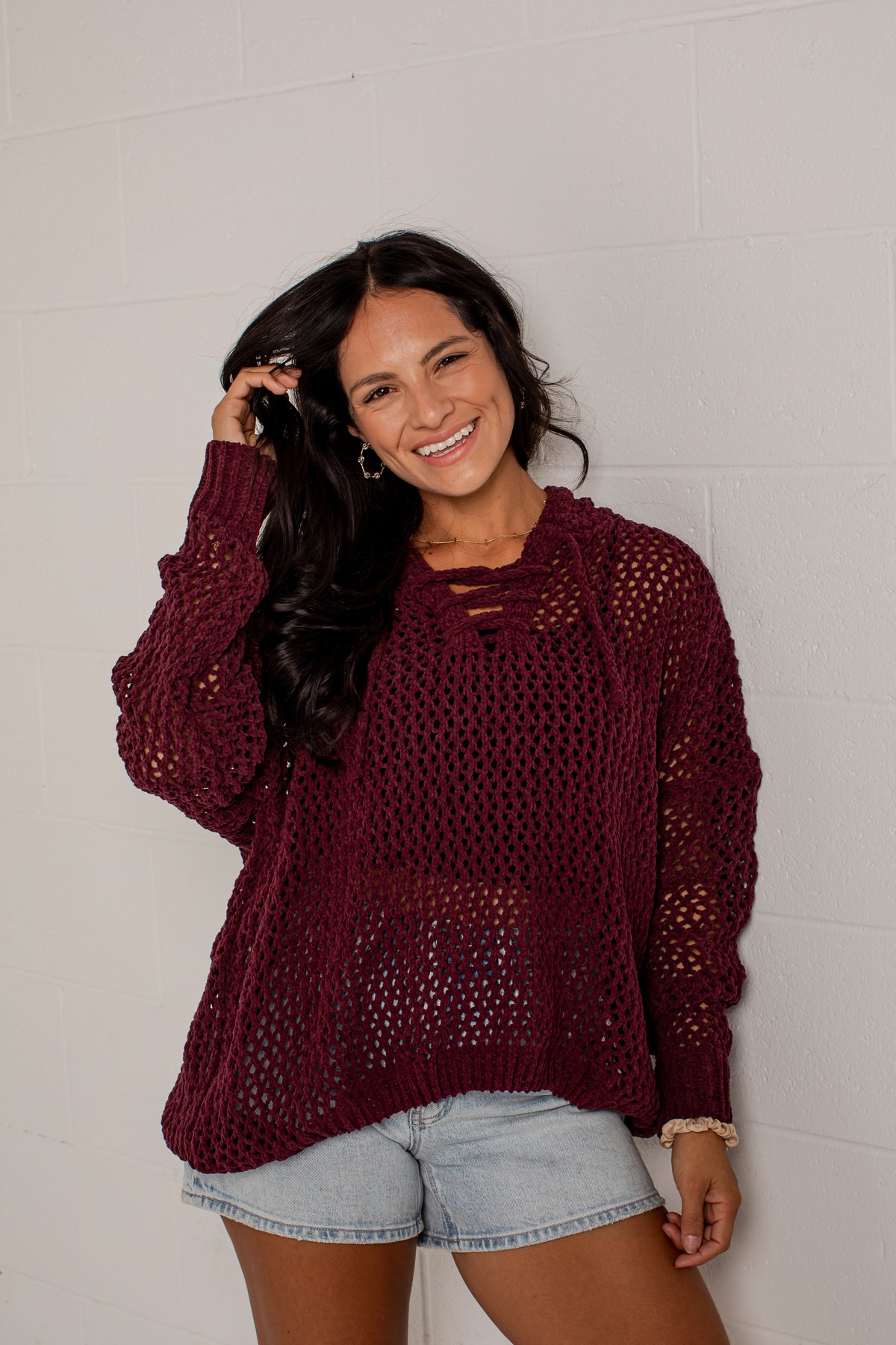 A woman wearing a maroon open-knit long-sleeve sweater and denim shorts, posing against a white brick wall.
