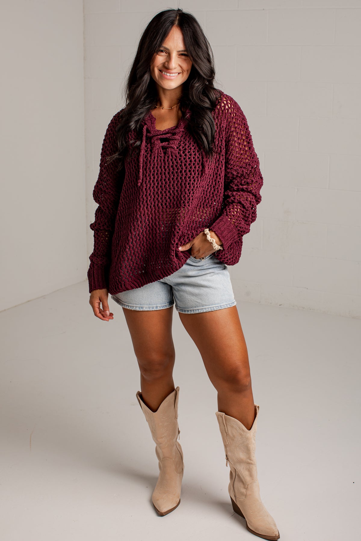 A woman modeling a chunky knit long-sleeve sweater in deep burgundy, paired with light-wash denim shorts and beige cowboy boots, standing against a neutral studio backdrop.