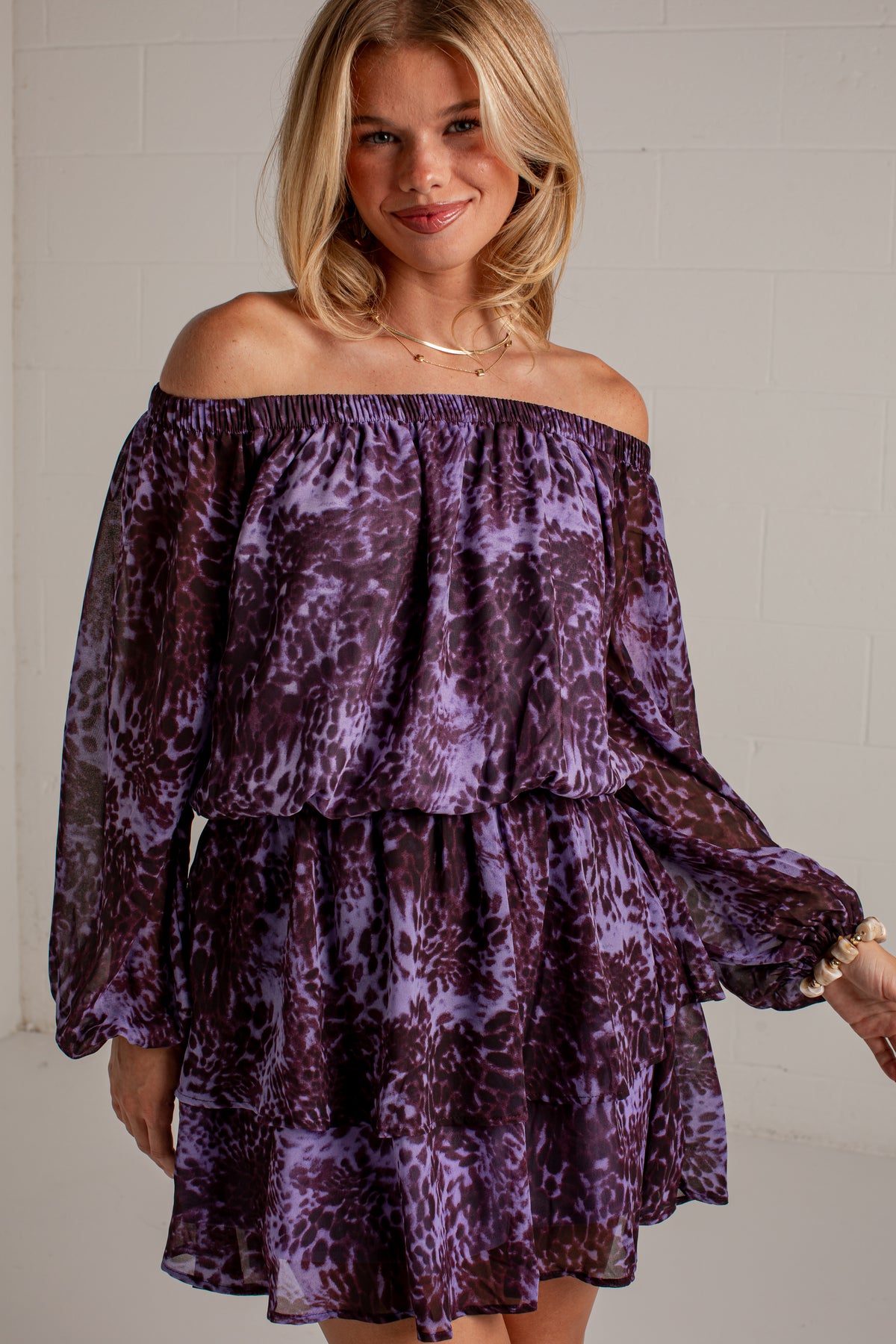 A woman wearing a purple and black tie-dye off-the-shoulder mini dress with long balloon sleeves, shown against a white brick wall.