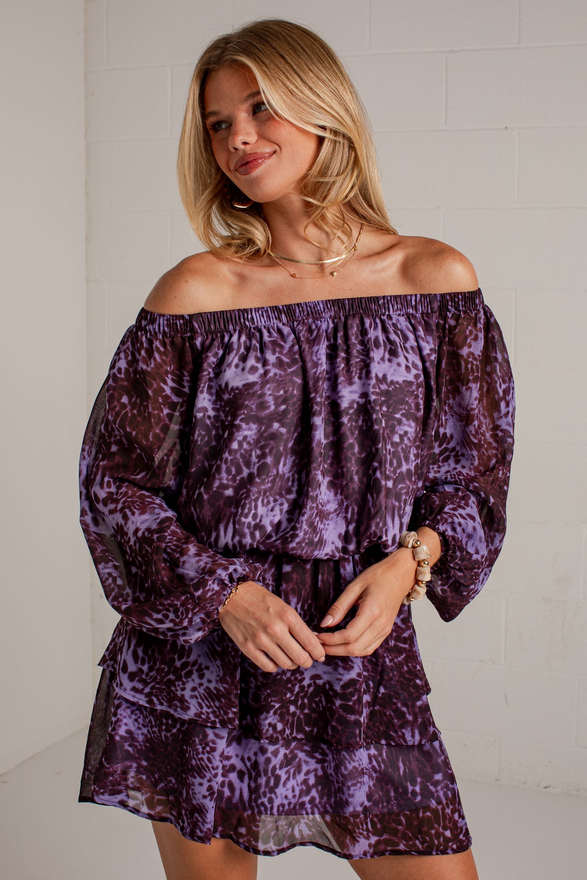 A woman wearing a purple off-the-shoulder long-sleeve mini dress with an abstract animal print pattern, standing against a white brick wall.