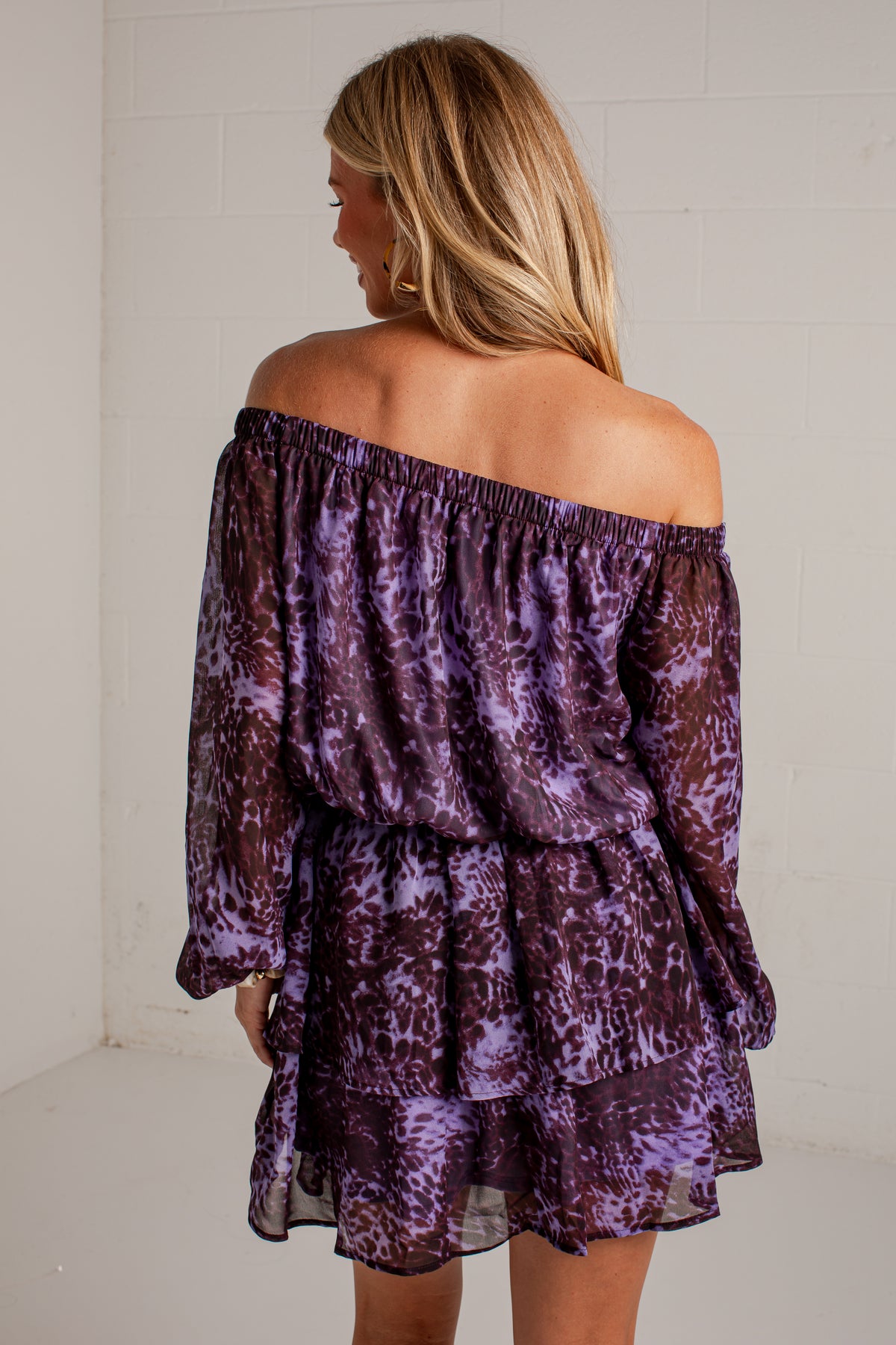 A woman shown from the back wearing a purple and dark brown abstract patterned off-the-shoulder dress with long sleeves and a tiered ruffle hem, standing against a white brick wall.