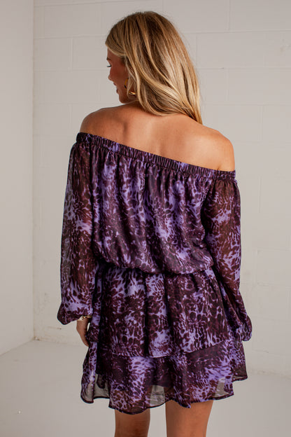 A woman shown from the back wearing a purple and dark brown abstract patterned off-the-shoulder dress with long sleeves and a tiered ruffle hem, standing against a white brick wall.