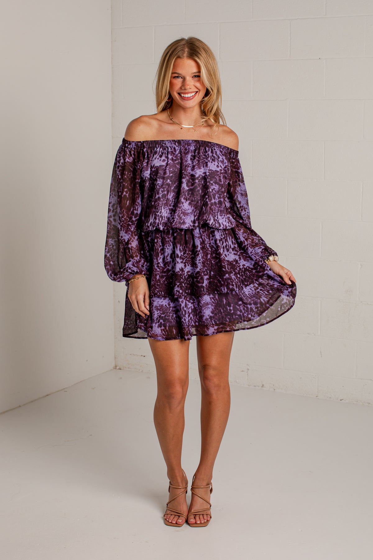 A woman modeling an off-the-shoulder purple tiered mini dress with a textured, crushed velvet-like pattern, paired with brown strappy high-heeled sandals, standing against a white brick wall.