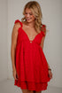 A woman wearing a bright red eyelet mini dress with ruffled shoulder straps and a keyhole cutout at the bust, standing against a plain off-white wall.