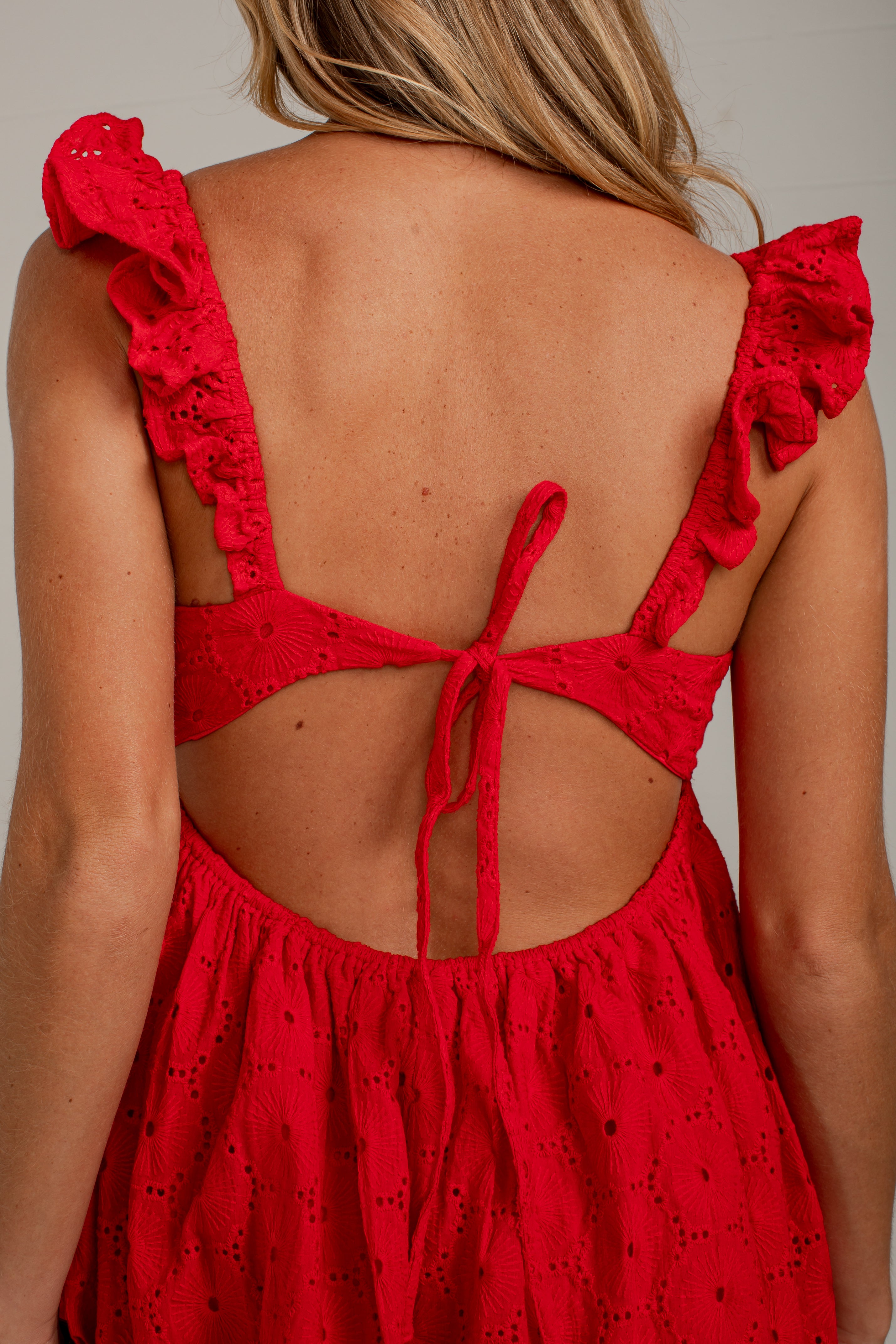 Close-up back view of a woman wearing a bright red eyelet dress with ruffled shoulder straps and a tie-back neckline, shown against a plain light gray background.