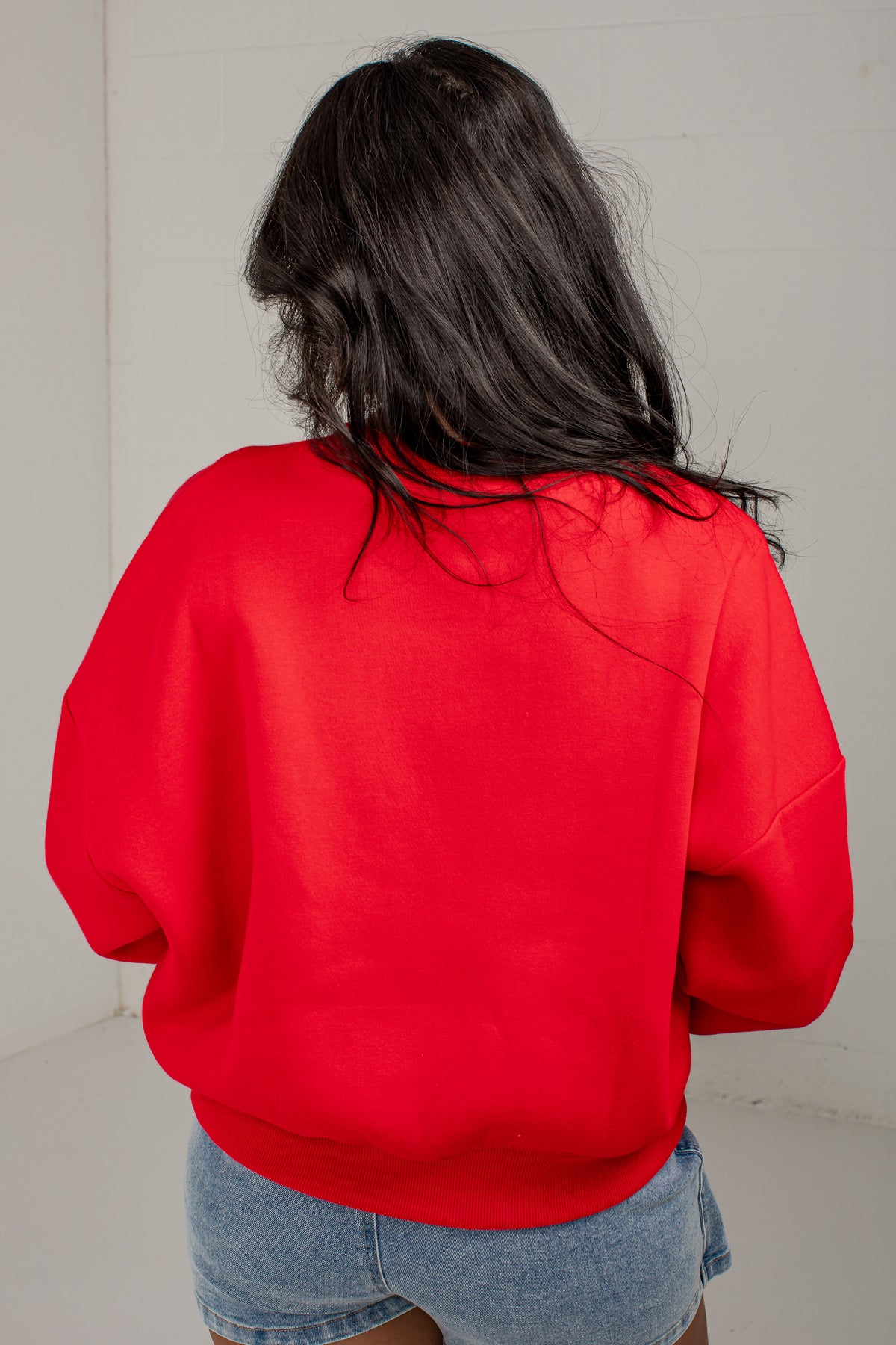 A person viewed from behind wearing a bright red crewneck sweatshirt and light blue denim shorts against a plain off-white wall.