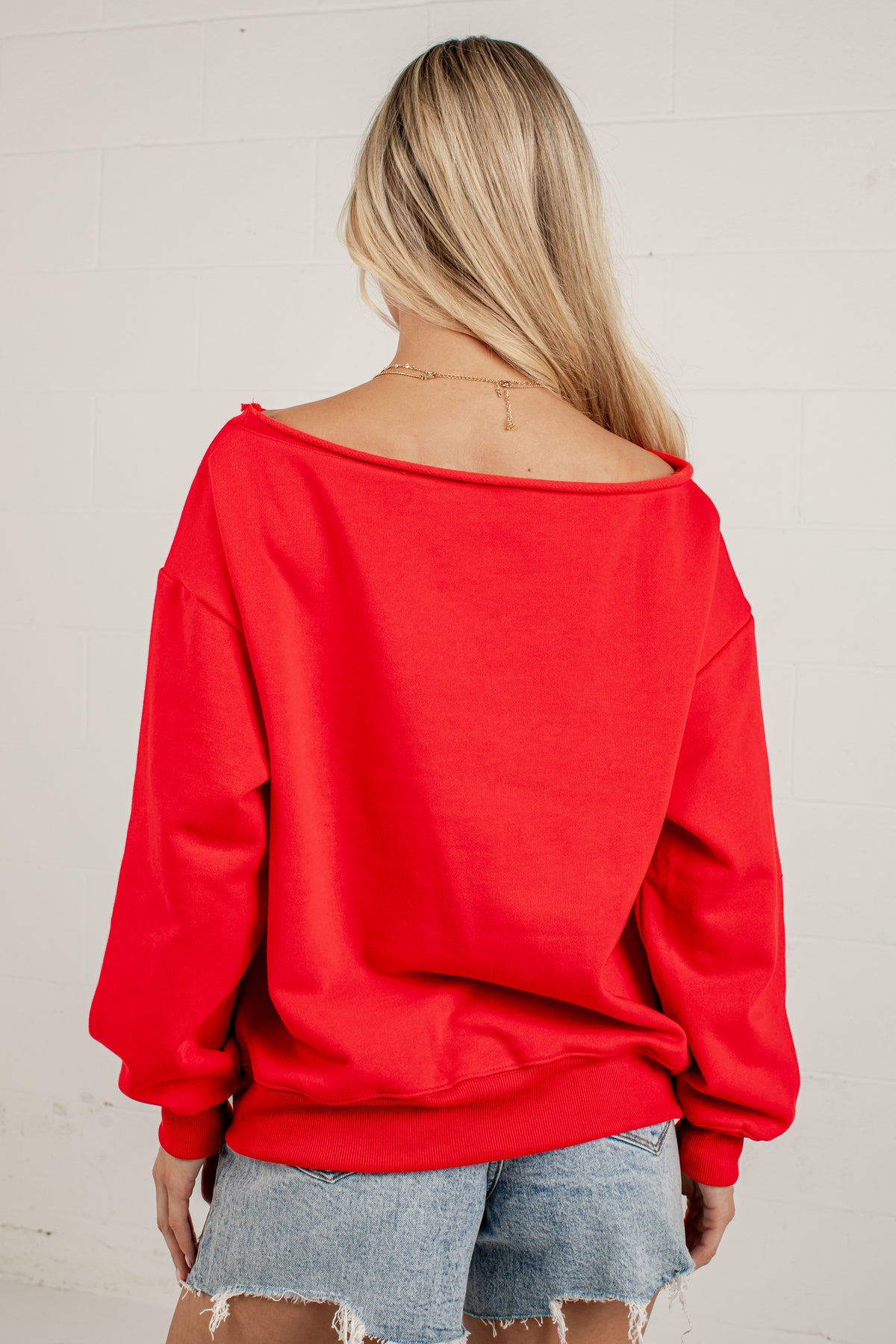 A woman viewed from behind wearing a bright red off-the-shoulder sweatshirt and light blue denim shorts against a white brick wall.