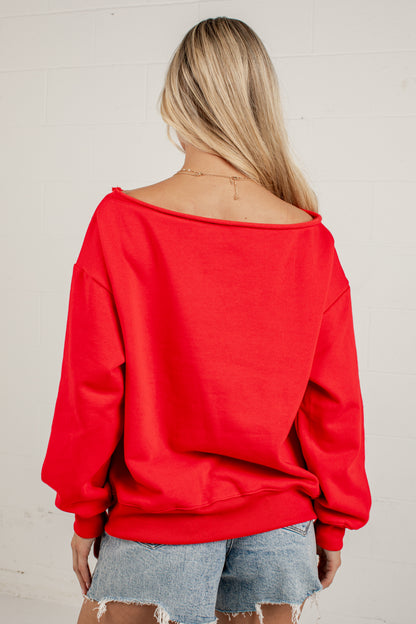 A woman viewed from behind wearing a bright red off-the-shoulder sweatshirt and light blue denim shorts against a white brick wall.