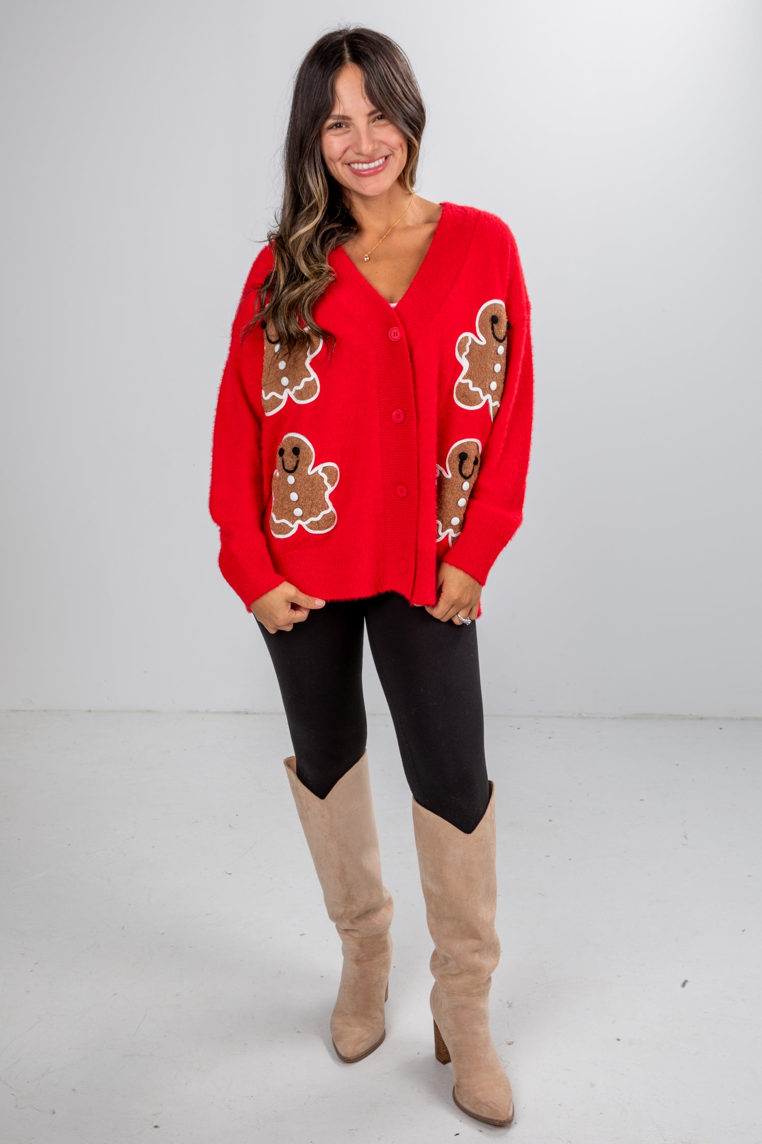 Woman wearing a red sweater with gingerbread man patterns, black leggings, and beige knee-high boots on a white background.