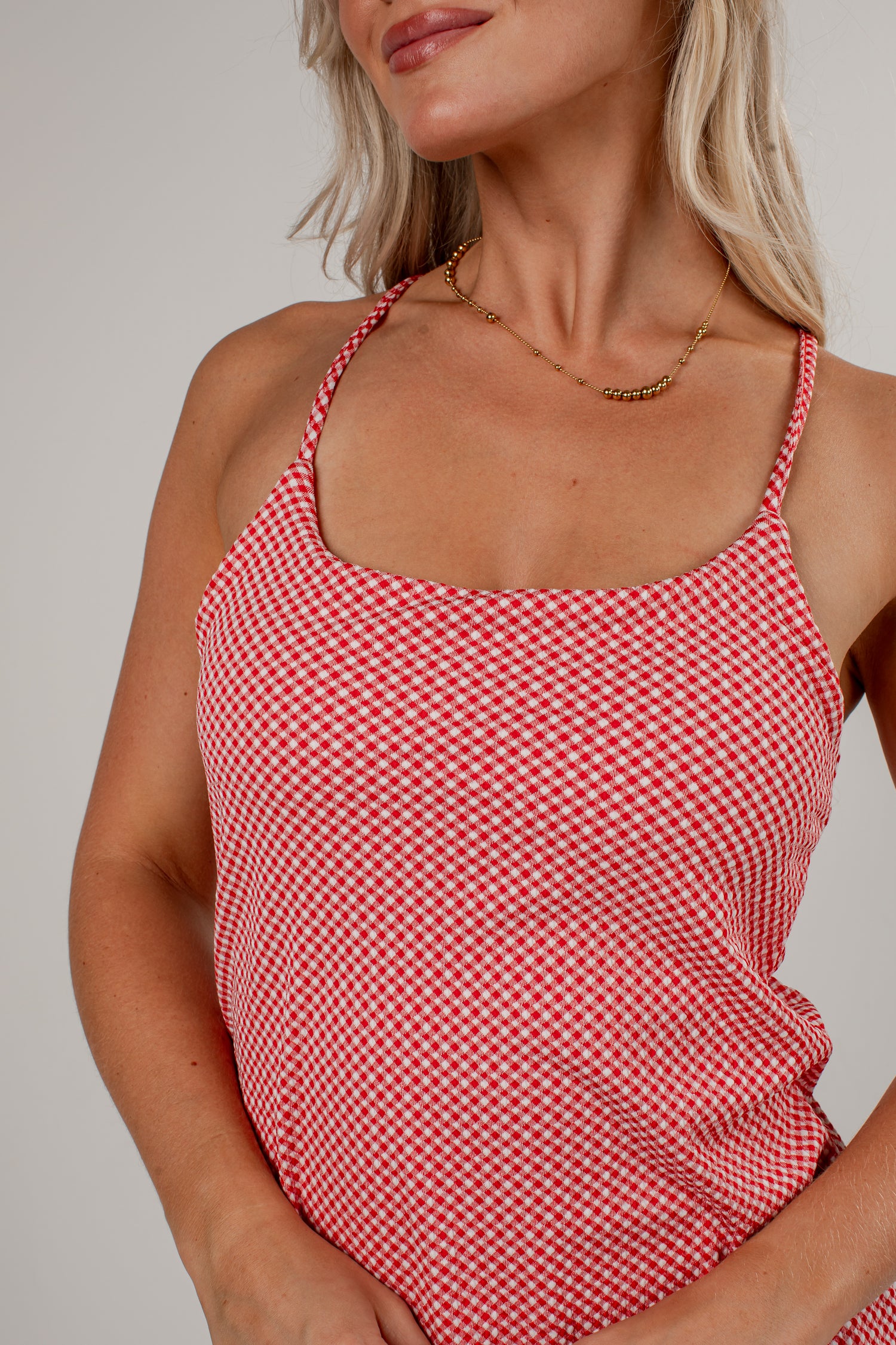 A close-up of a woman wearing a red and white gingham patterned camisole top with thin straps, paired with a delicate gold chain necklace, against a neutral light gray background.