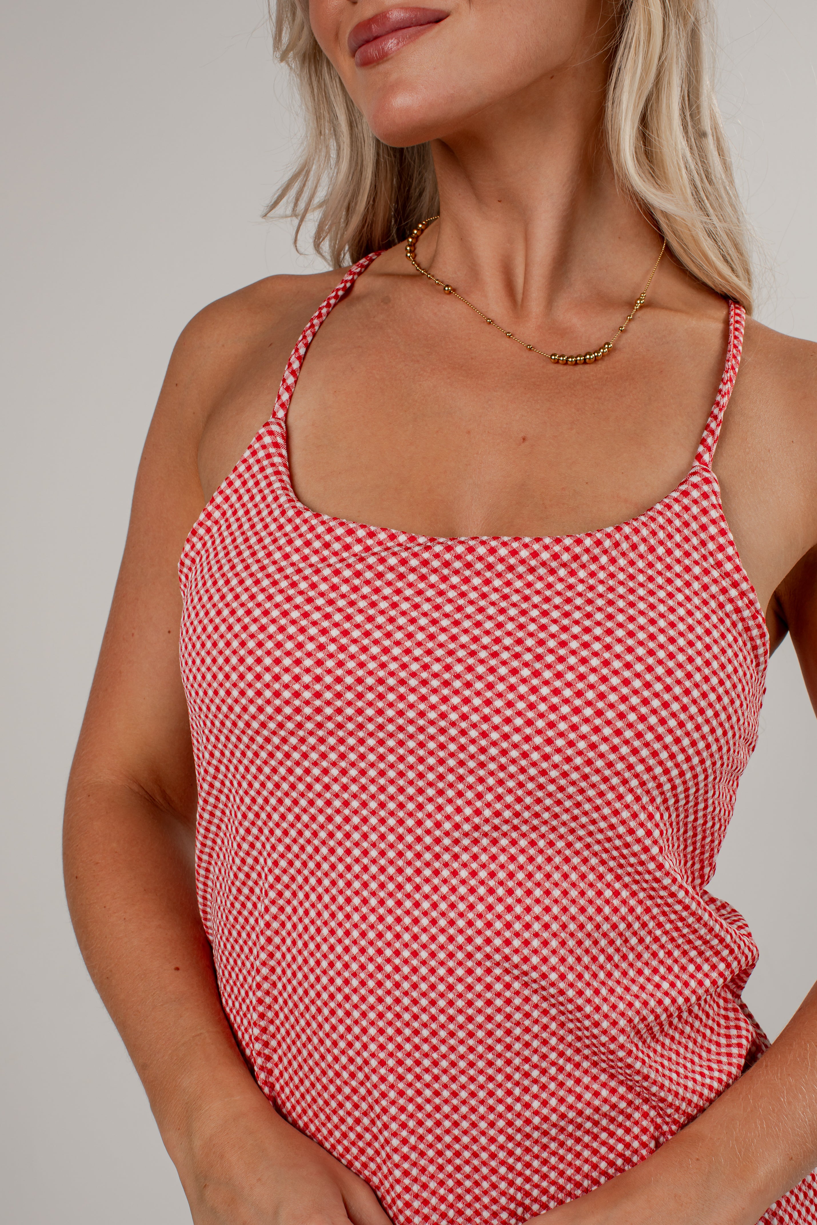 A close-up of a woman wearing a red and white gingham patterned camisole top with thin straps, paired with a delicate gold chain necklace, against a neutral light gray background.
