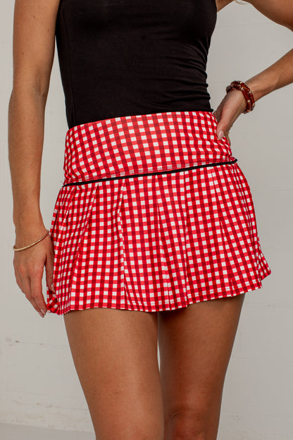 A close-up of a model wearing a red and white gingham pleated tennis skirt with a black waistband trim, paired with a black sleeveless top.